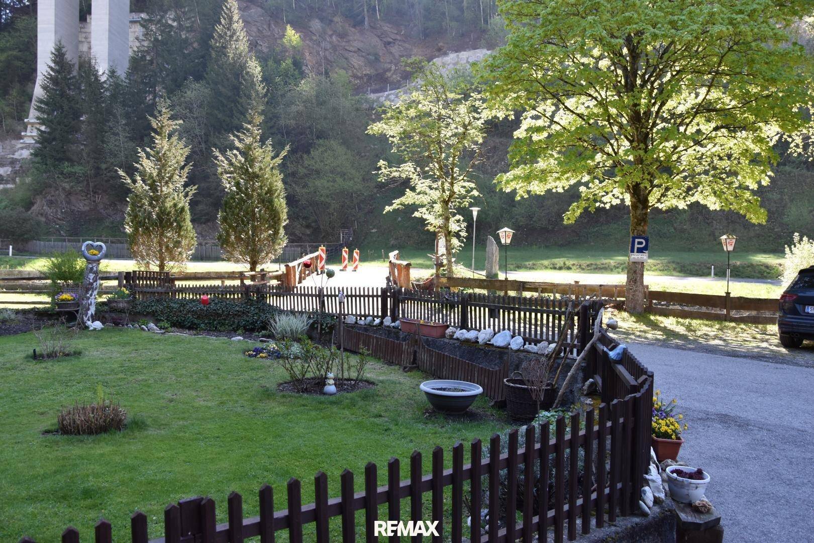 Gepflegter Gartenbereich mit Holzzaun, Rasenfläche und Bäumen, im Hintergrund eine Brückenkonstruktion.