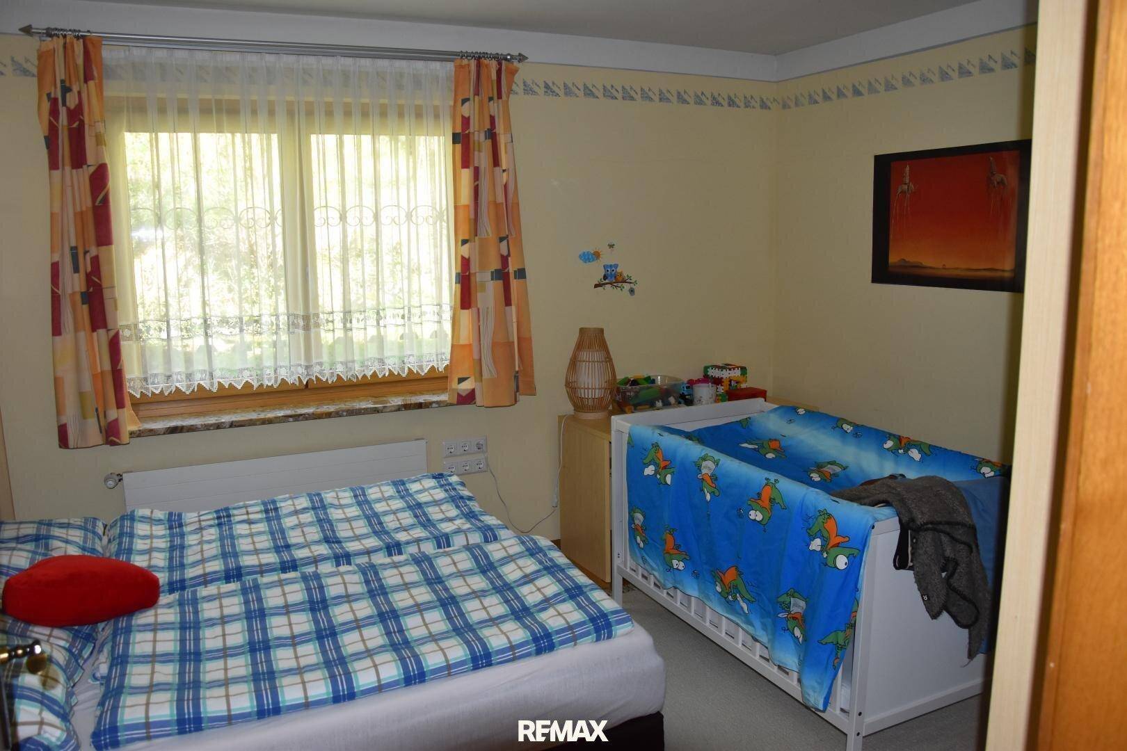 Schlafzimmer mit Doppelbett und Babybett, hell durch ein großes Fenster mit Vorhängen.