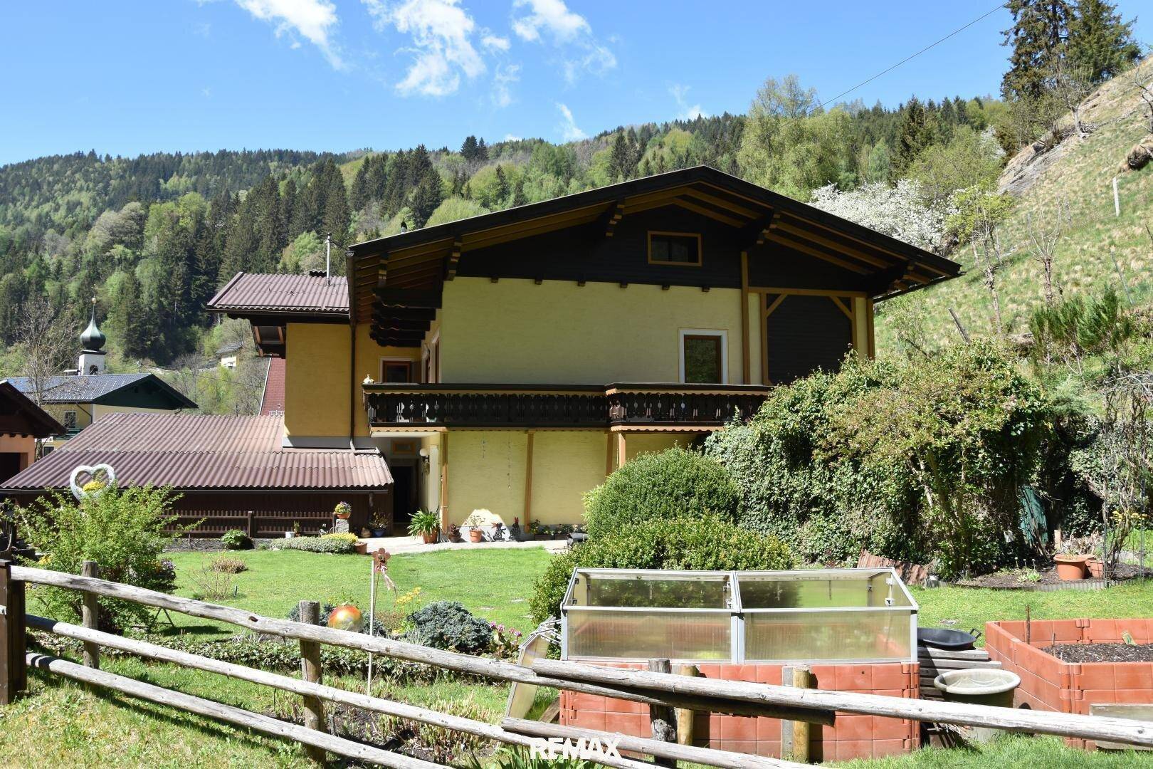 Hausansicht vom Garten aus mit Gewächshaus, Hochbeeten und grüner Umgebung.