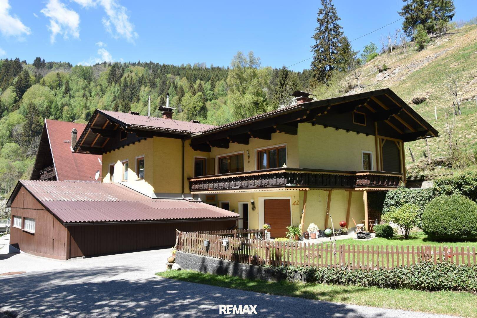 Traditionelles gelbes Haus mit Holzbalkon, Garage und gepflegtem Vorgarten in ländlicher Umgebung.