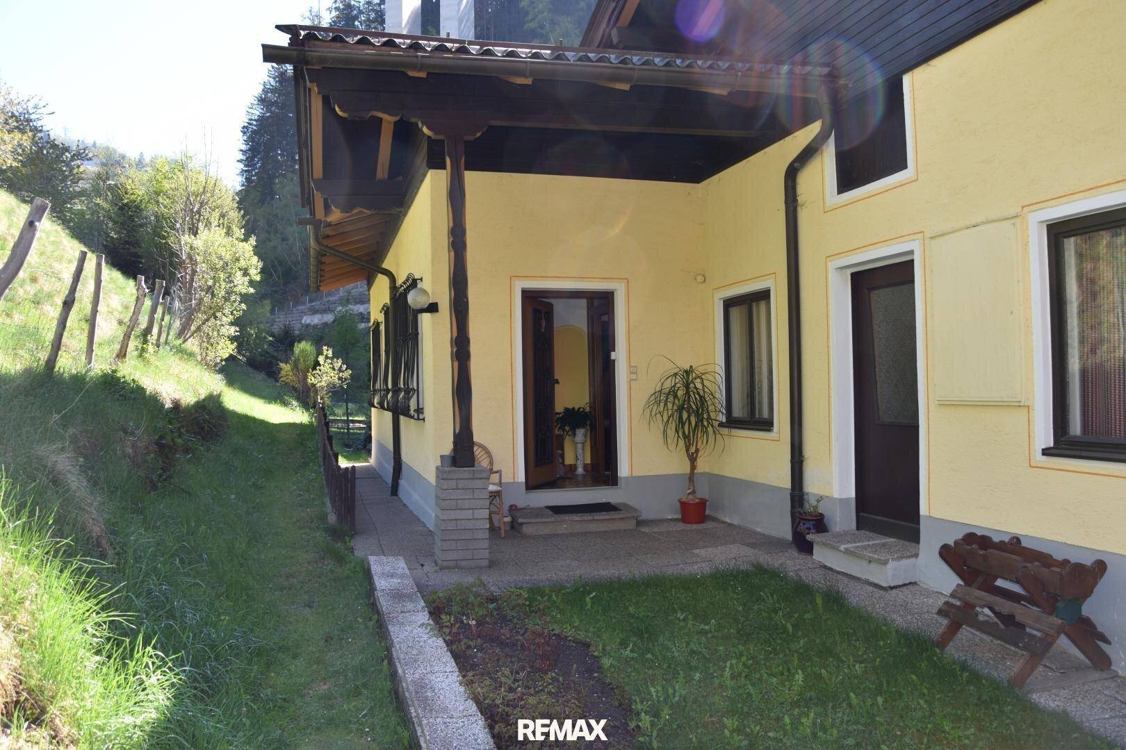 Seitlicher Eingangsbereich des Hauses mit überdachtem Vorbau und angrenzendem grünen Hang.