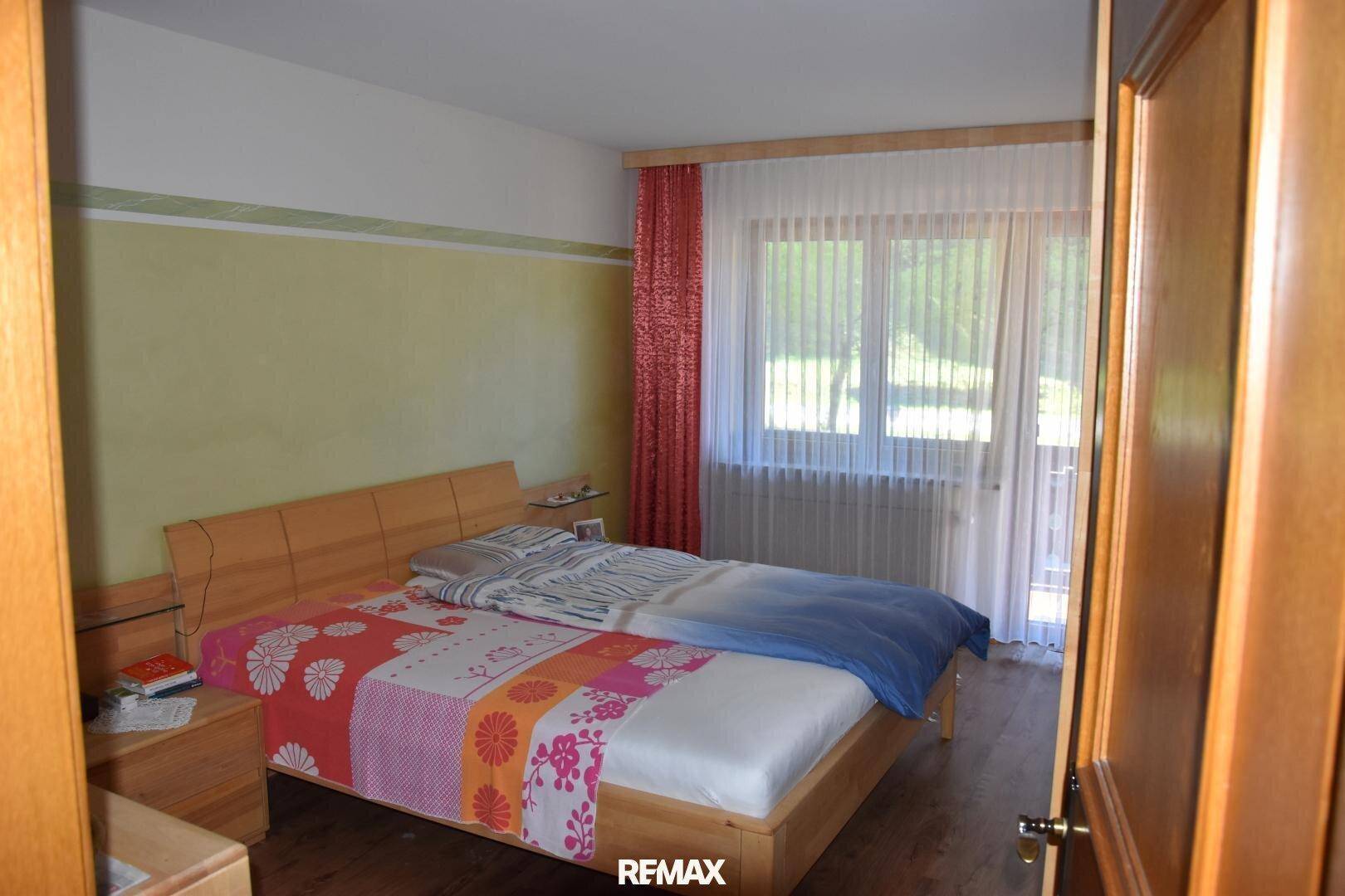 Schlafzimmer mit Holzdoppelbett, Holzboden und Zugang zu einem Balkon durch ein großes Fenster.