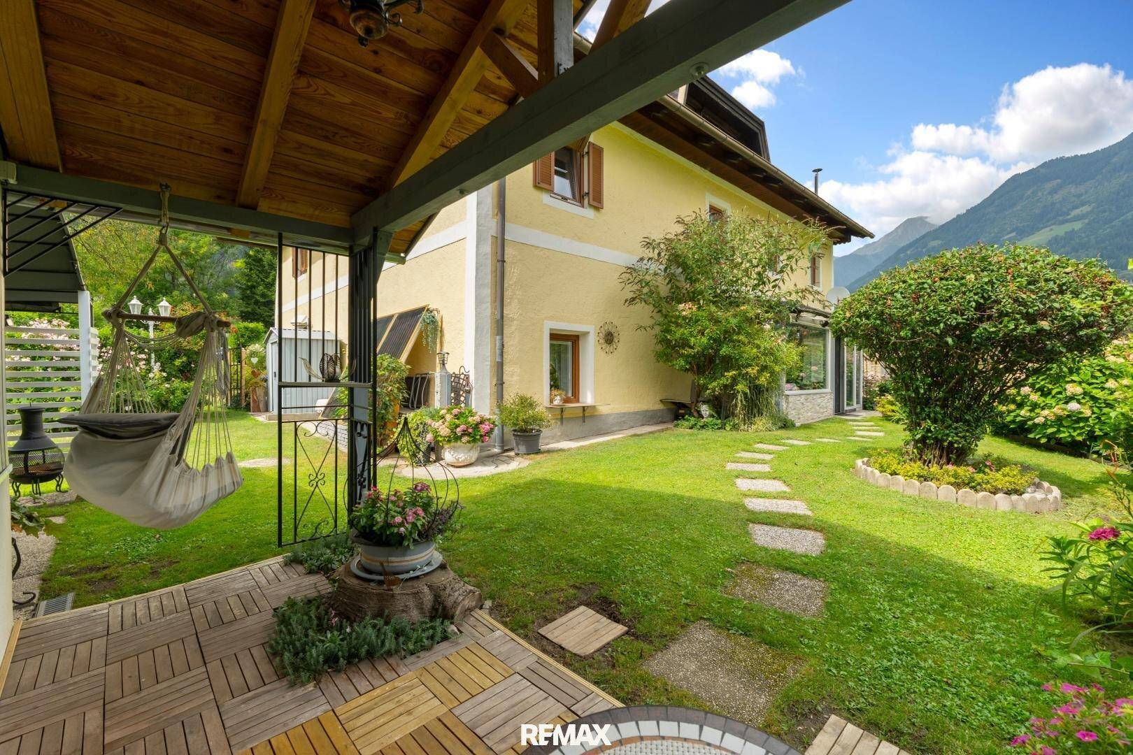 Sonniger Gartenbereich mit Holzterrasse, Hängematte und Blick auf das Haus und die umliegende Natur.