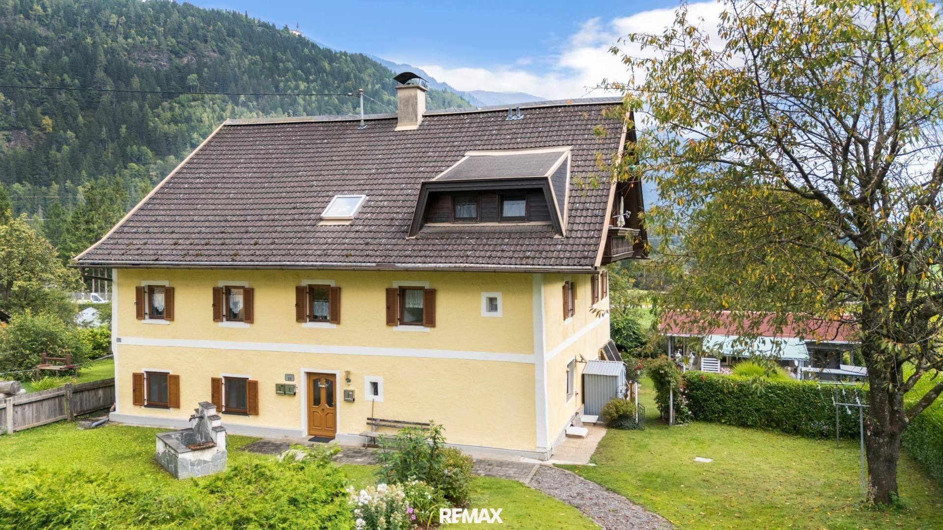 Traditionelles gelbes Haus mit Satteldach, umgeben von einem grünen Garten und Berglandschaft.