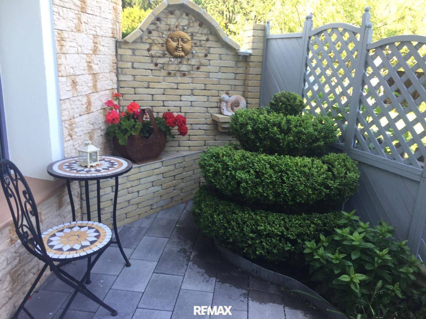 Kleiner gepflasterter Patio mit dekorativer Steinwand, Sitzgelegenheit und gepflegten Sträuchen.