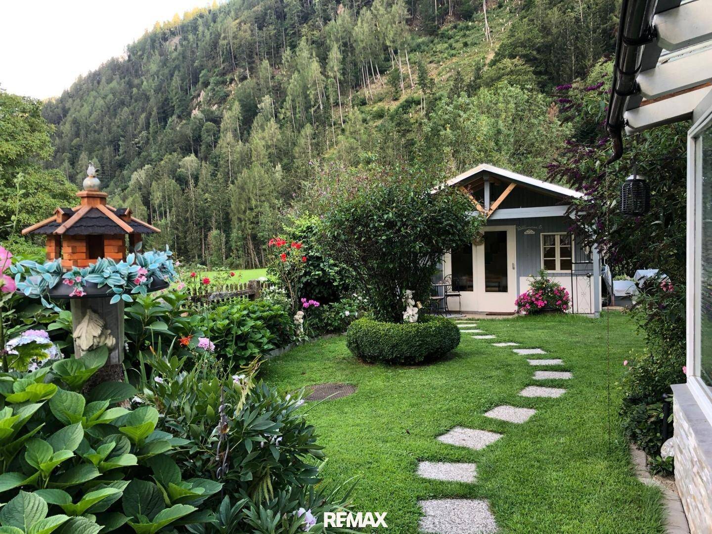 Gepflegter Garten mit Steinpfad, üppiger Bepflanzung und einem charmanten Gartenhaus vor Bergkulisse.