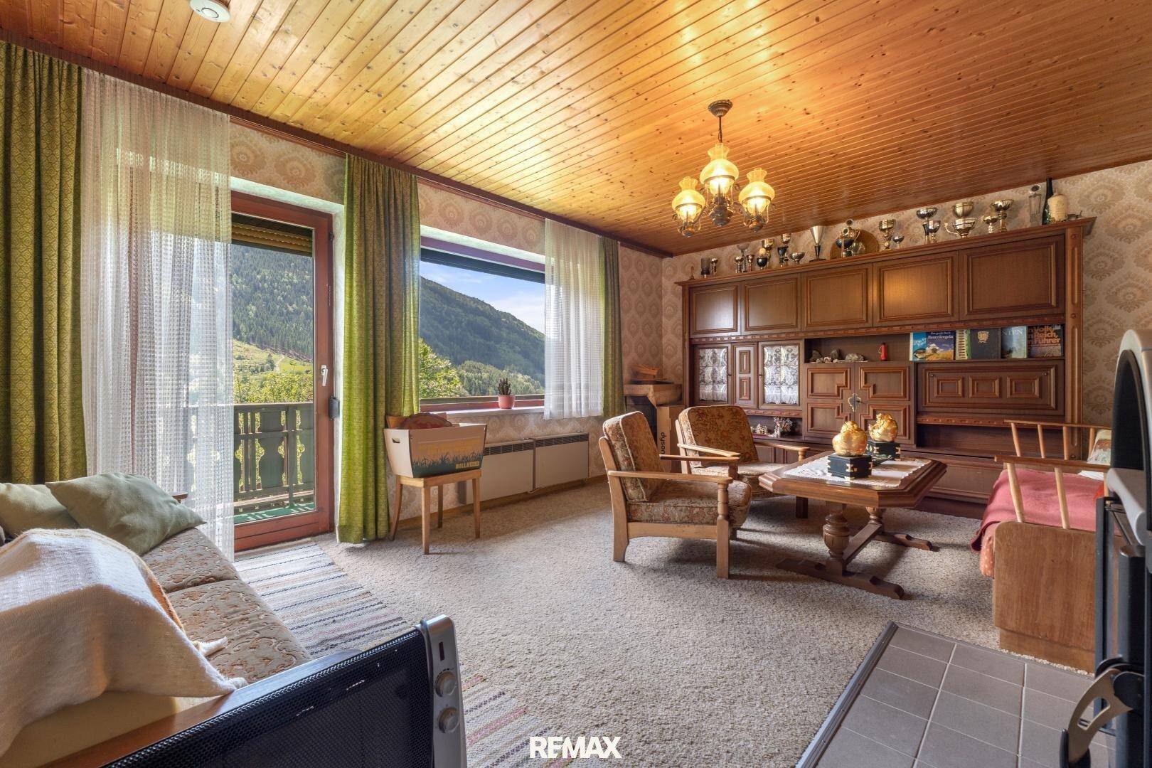 Gemütliches Wohnzimmer im alpinen Stil mit Holzdecke, Teppichboden und Bergblick vom Balkon.