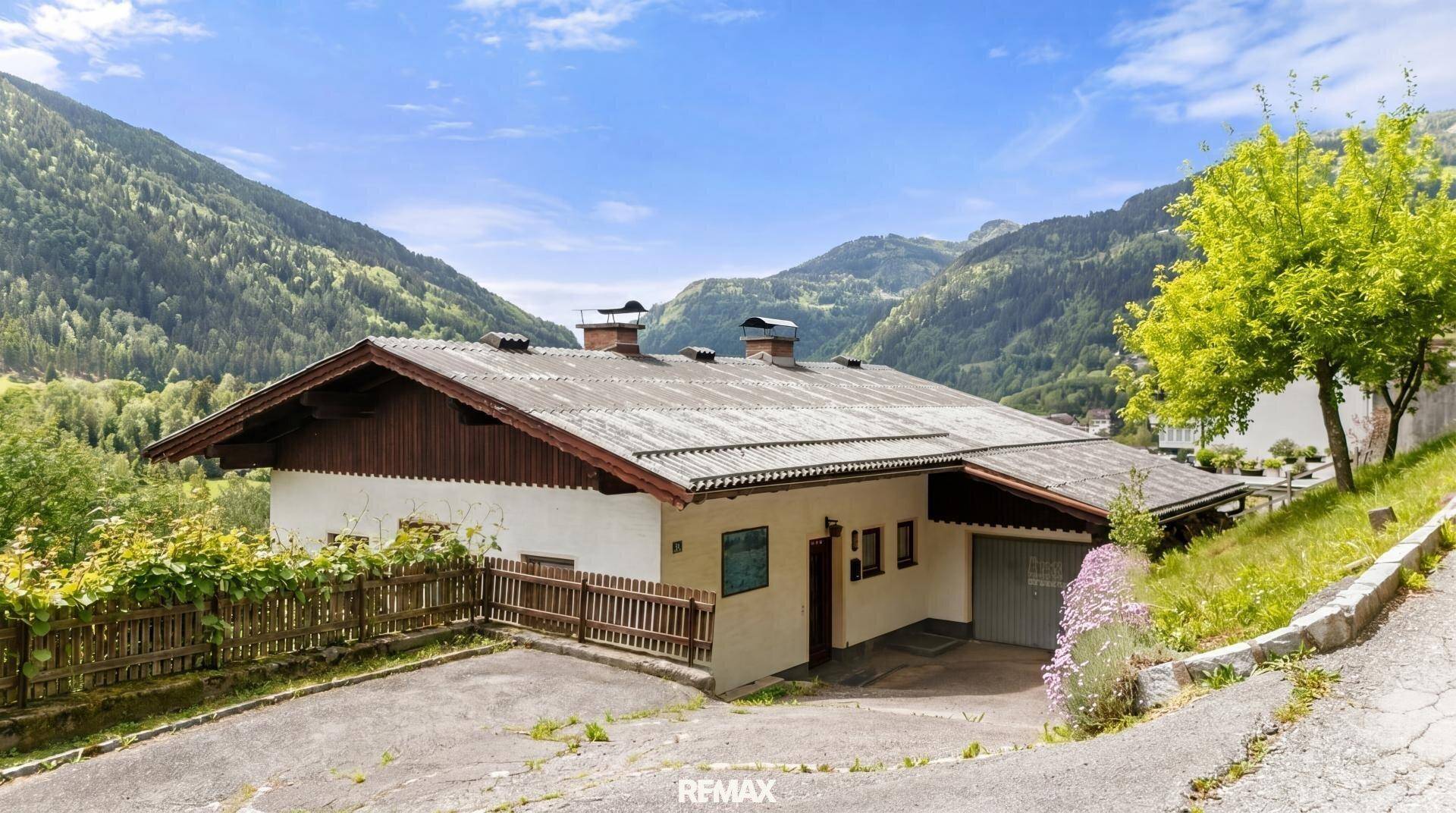 Vorderansicht des Hauses mit Garage, umgeben von üppiger Vegetation und Bergpanorama.