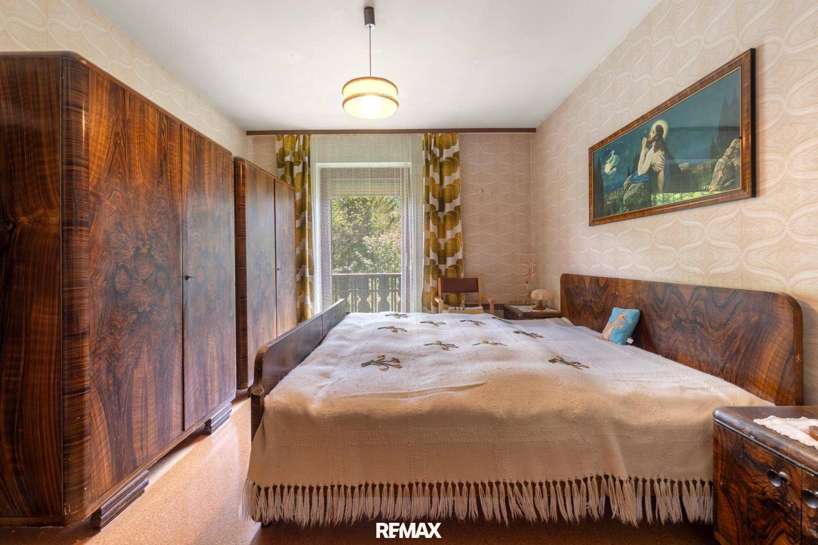 Schlafzimmer mit Vintage-Möbeln, gemusterter Tapete und Balkonzugang mit Blick ins Grüne.