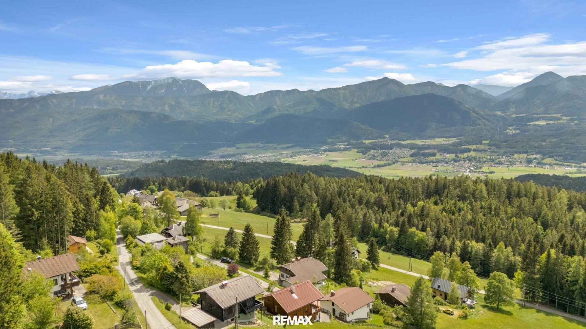 Panoramablick über eine ländliche Siedlung mit bewaldeten Hügeln und majestätischen Bergen im Hintergrund.