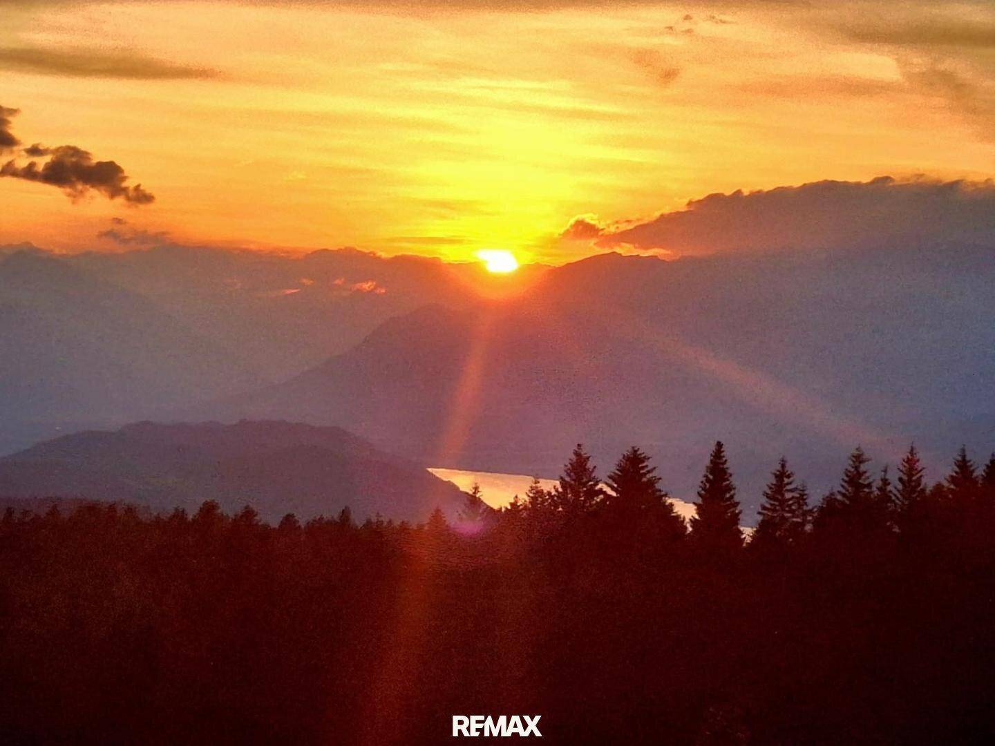 Spektakulärer Sonnenuntergang über einer Berglandschaft mit See und dunklen Wäldern im Vordergrund.