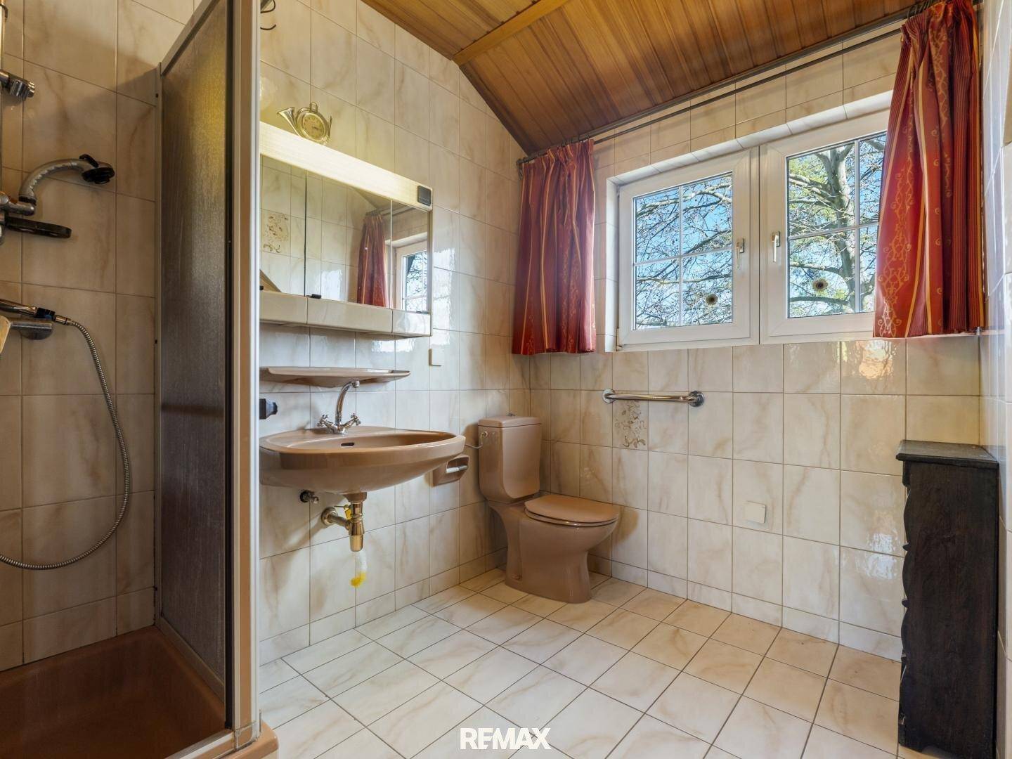 Badezimmer mit beigefarbenen Fliesen, Dusche, WC und Waschbecken unter einem Fenster.