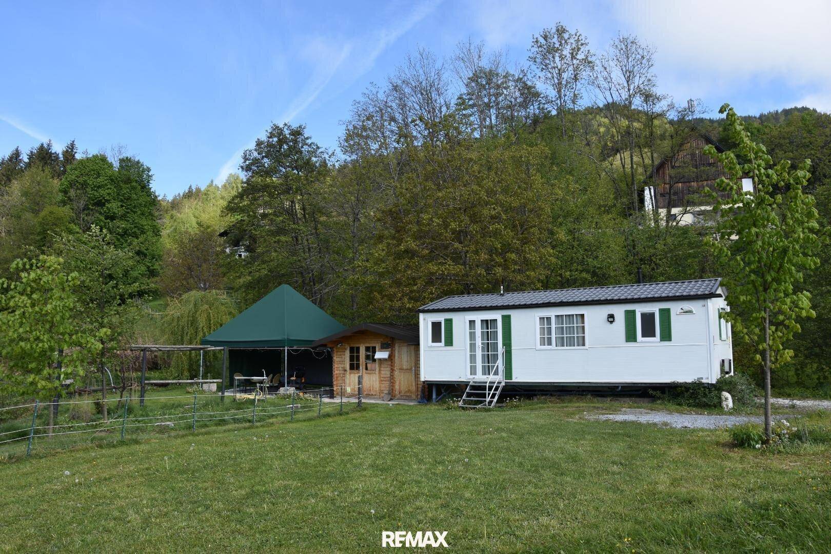 Mobilheim und Holzhütte in einem weitläufigen Gartenbereich mit grüner Umgebung und Hügeln.