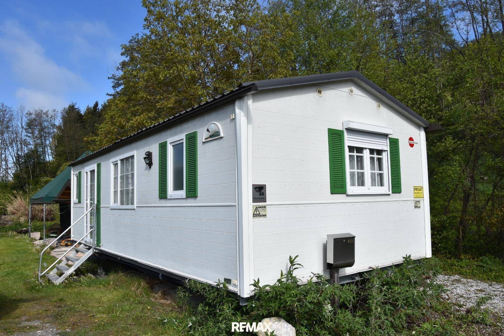 Weißes Mobilheim mit grünen Fensterläden und einer kleinen Treppe in ländlicher Umgebung.