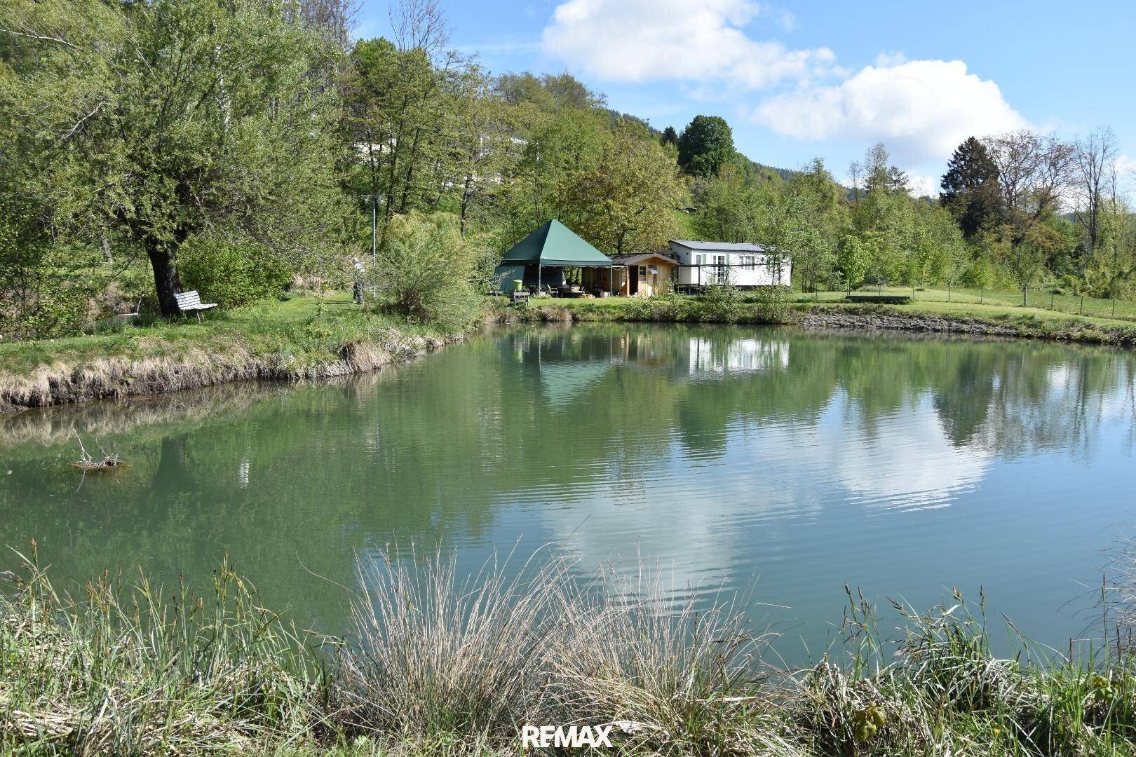 Malerischer Teich in einer weitläufigen Gartenlandschaft mit Mobilheim und Partyzelt im Hintergrund.