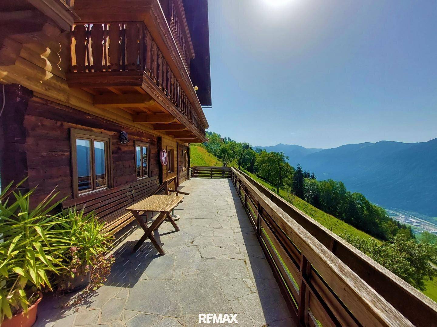 Lange, steingepflasterte Terrasse am Holzhaus mit Holzbänken und einem Tisch, bietet einen weiten Blick ins Tal und auf die Berge.