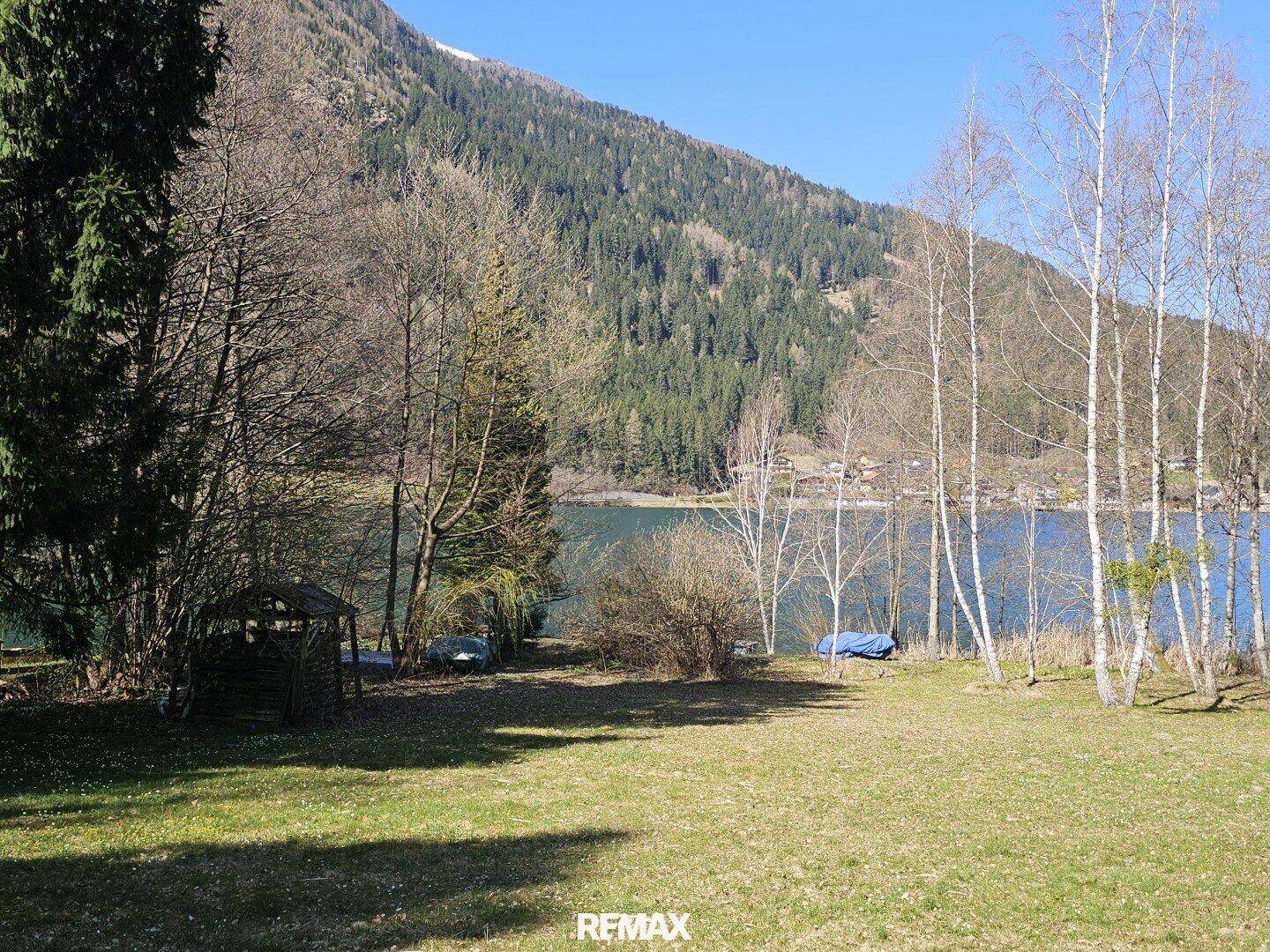Weitläufige Rasenfläche mit verschiedenen Bäumen und Blick auf den See und die Berge.