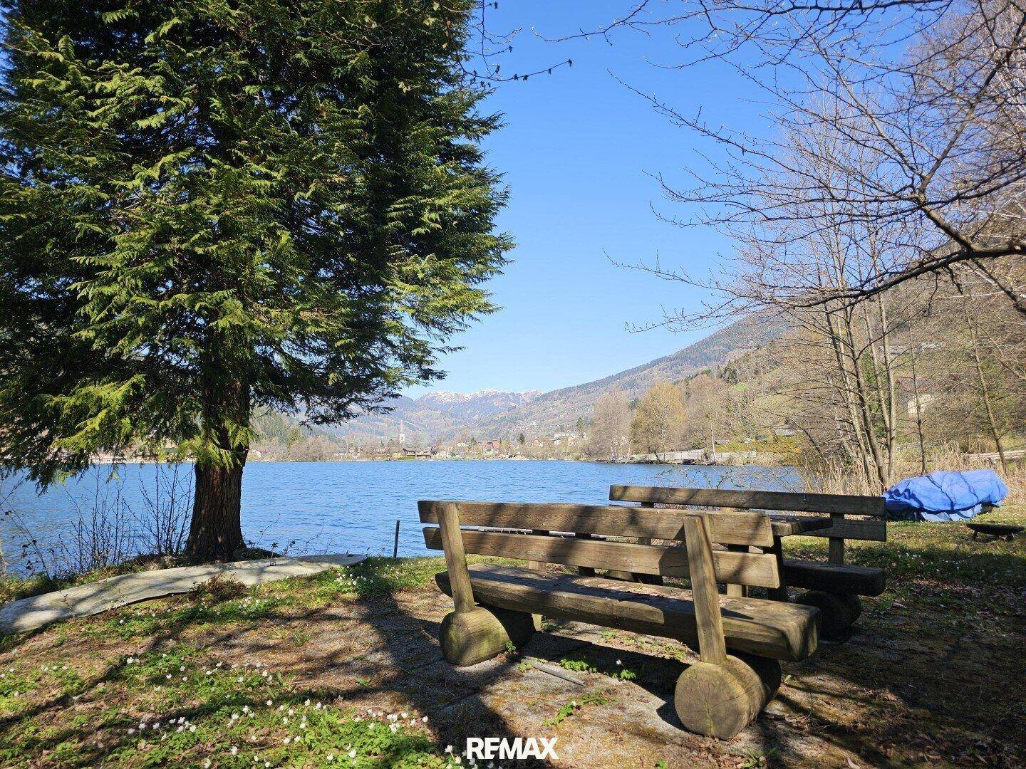 Rustikale Holzbänke am Seeufer mit weitem Blick auf ein Dorf und schneebedeckte Berge.