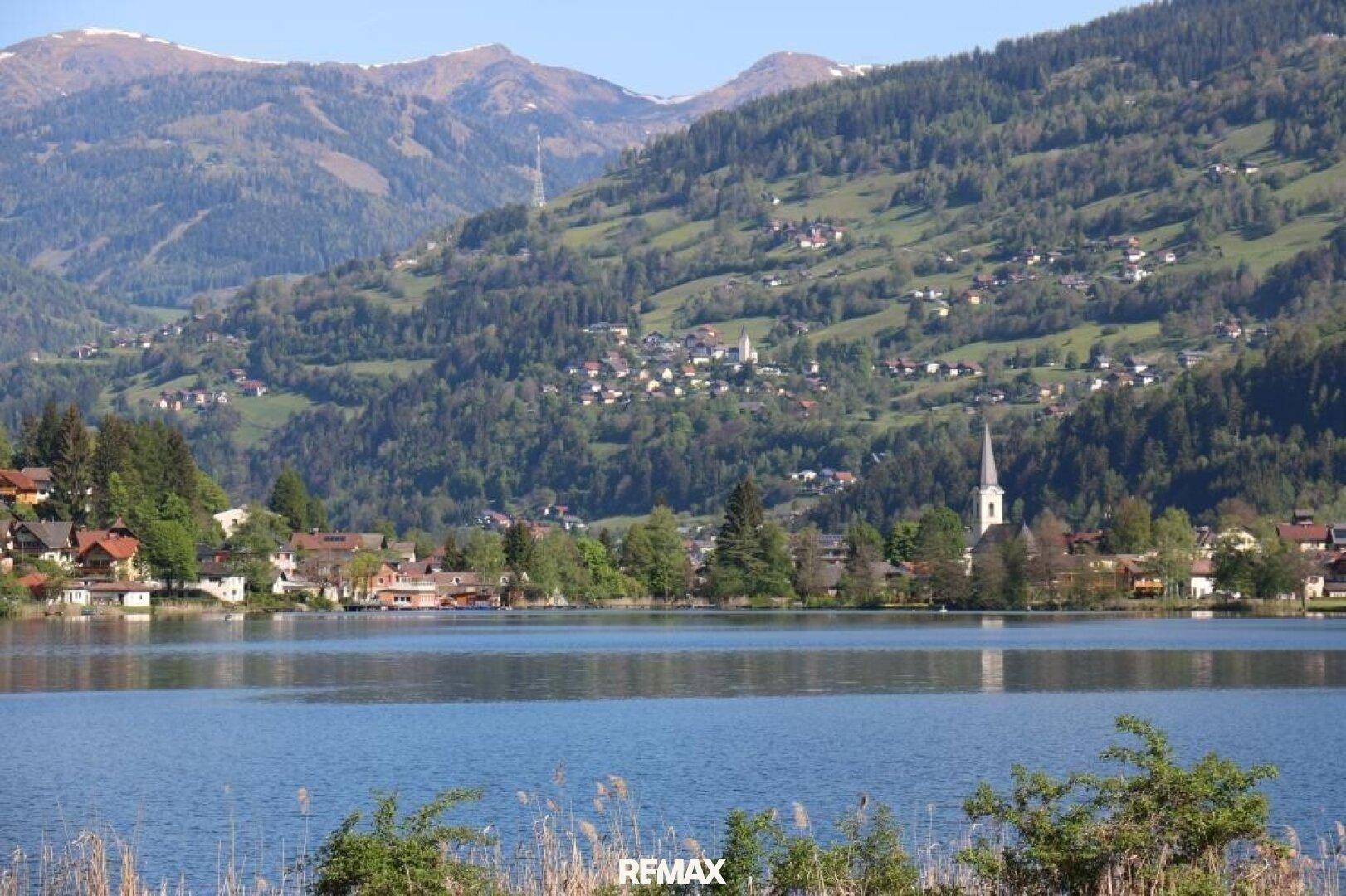 Panoramablick über einen See auf ein malerisches Dorf mit Kirche und bewaldeten Berghängen.