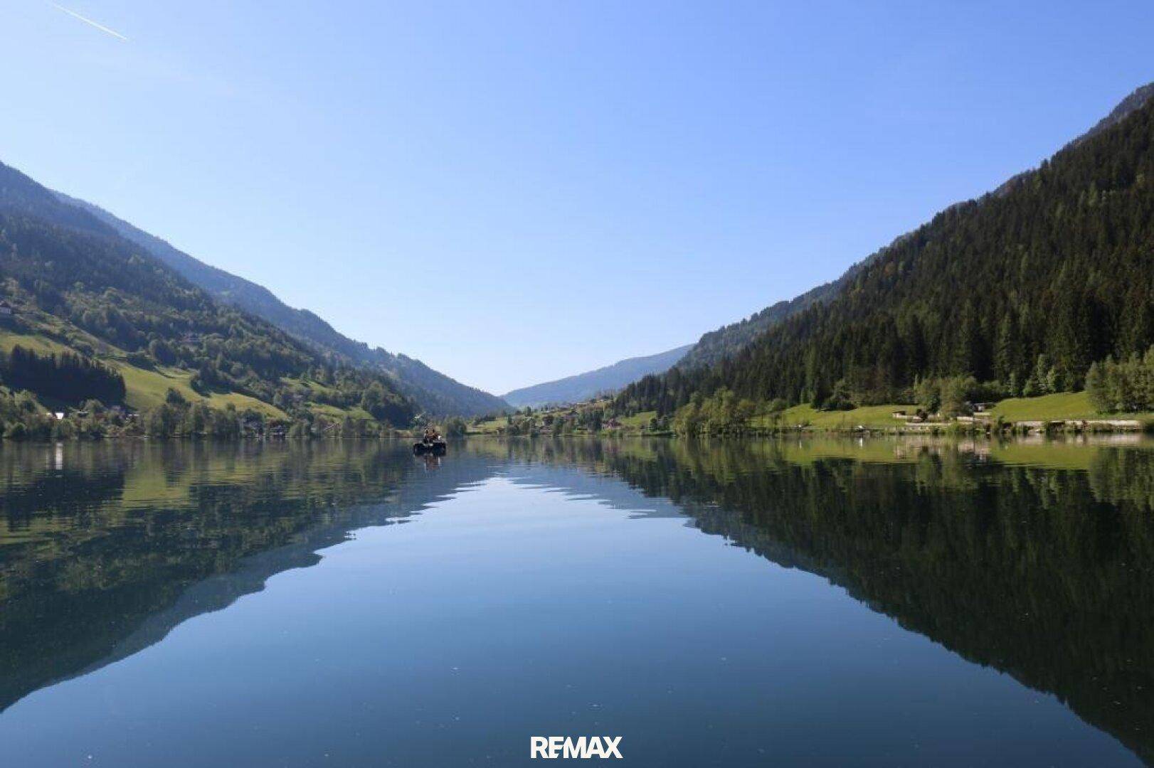 Weiter Blick über einen spiegelglatten See, umgeben von bewaldeten Bergen unter blauem Himmel.