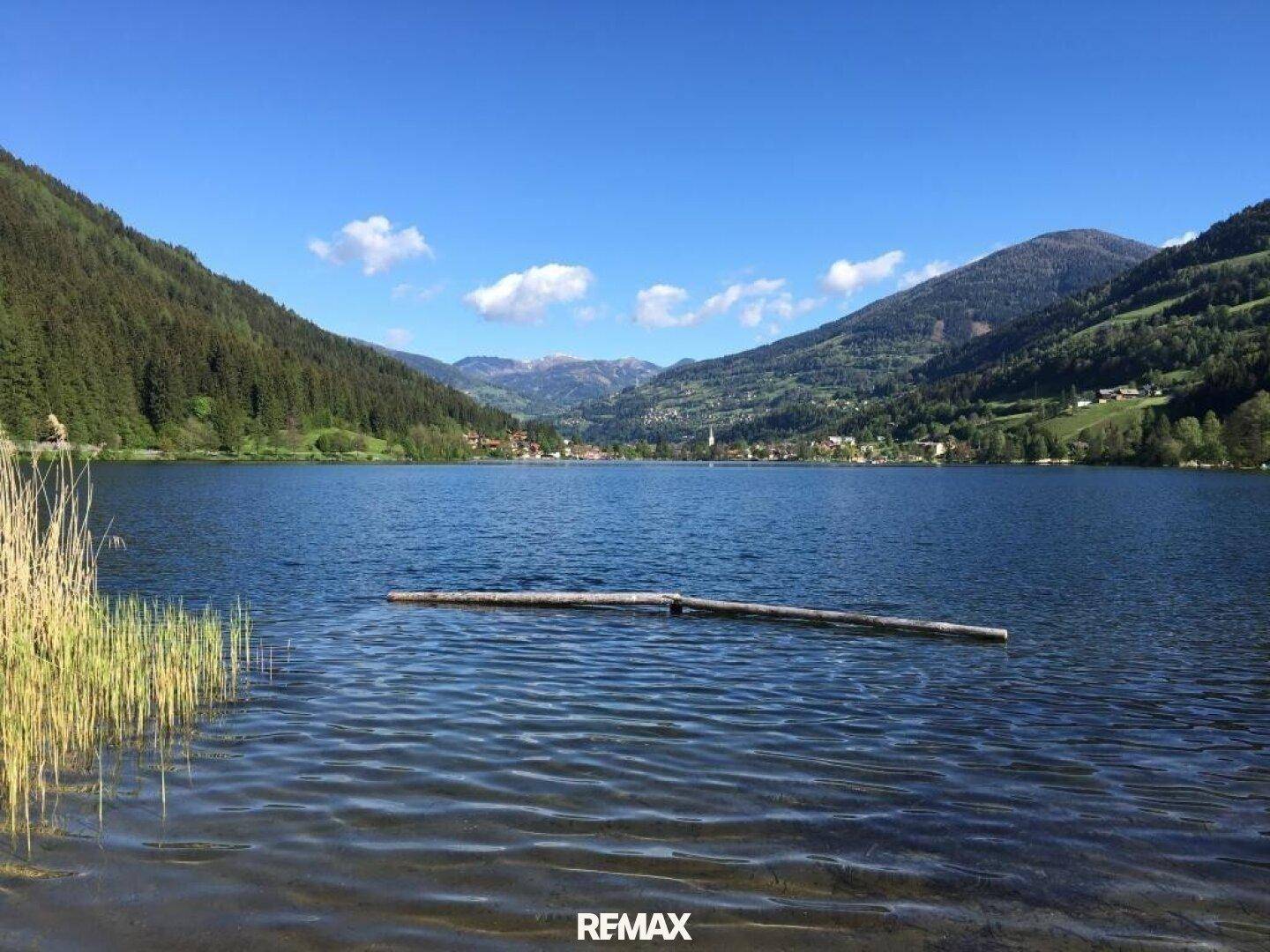 Breiter Blick über den See mit Schilf im Vordergrund und einem Dorf vor den Bergen.