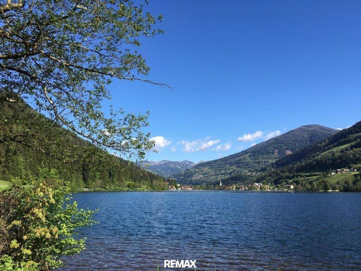 Blick auf den See durch Baumzweige hindurch, mit einem Dorf und Bergen in der Ferne.