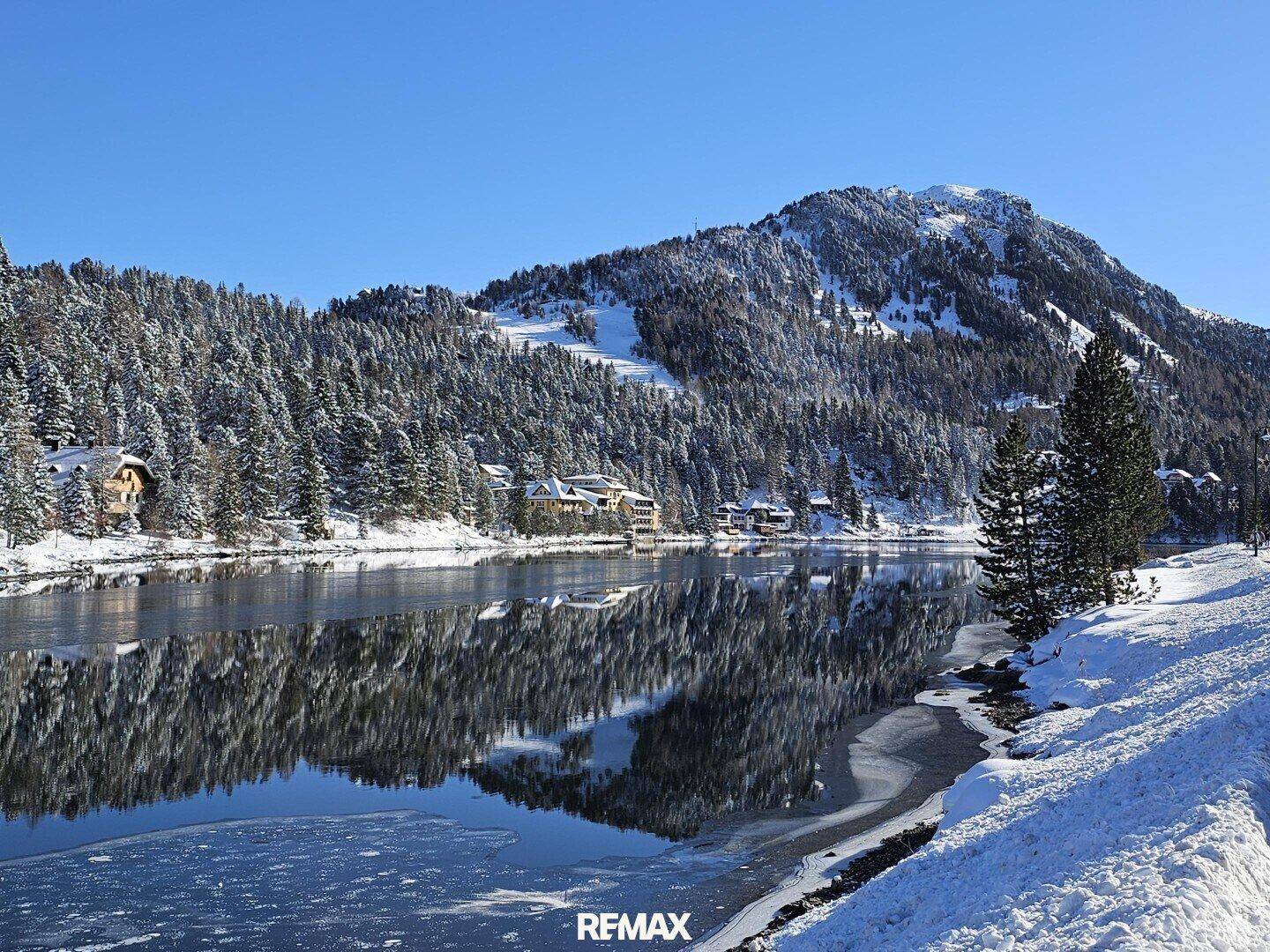Malerischer See mit teilweiser Eisdecke, umgeben von schneebedeckten Wäldern und Bergen.
