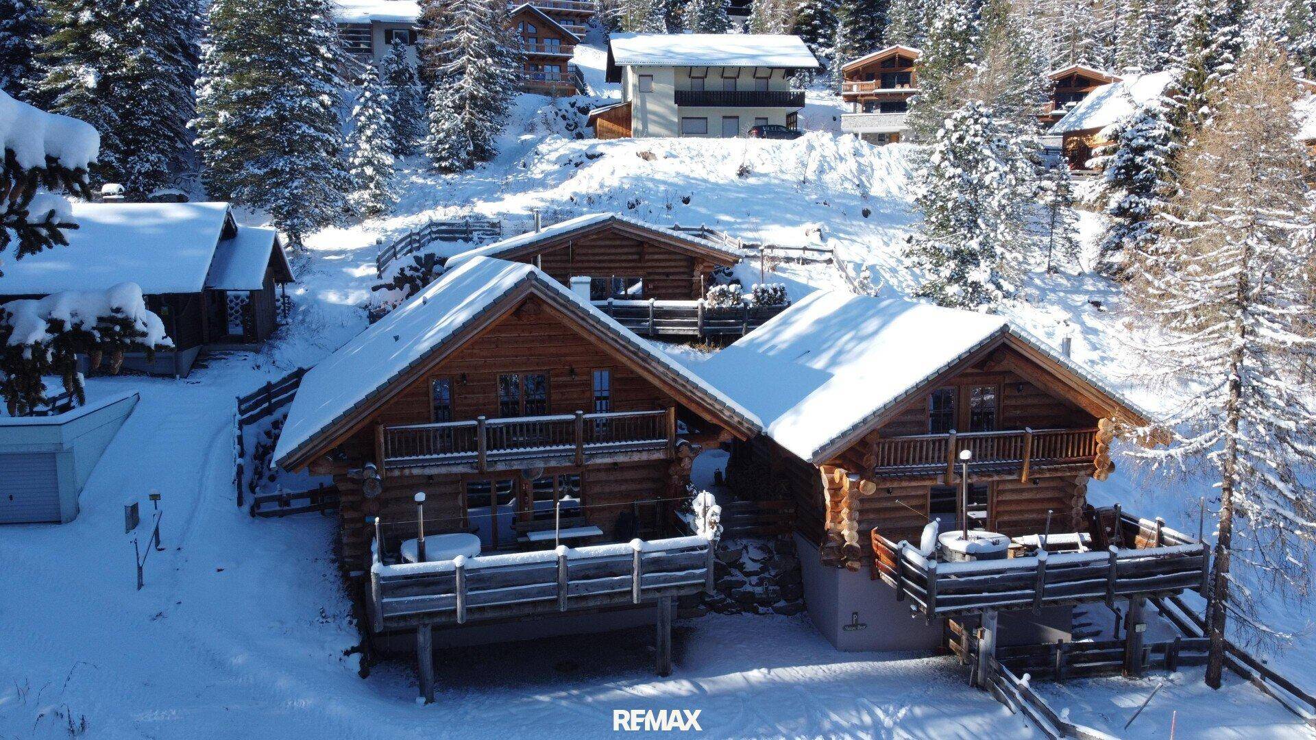 Luftaufnahme mehrerer Holzchalets in einer idyllischen, schneebedeckten Berglandschaft mit Wald.