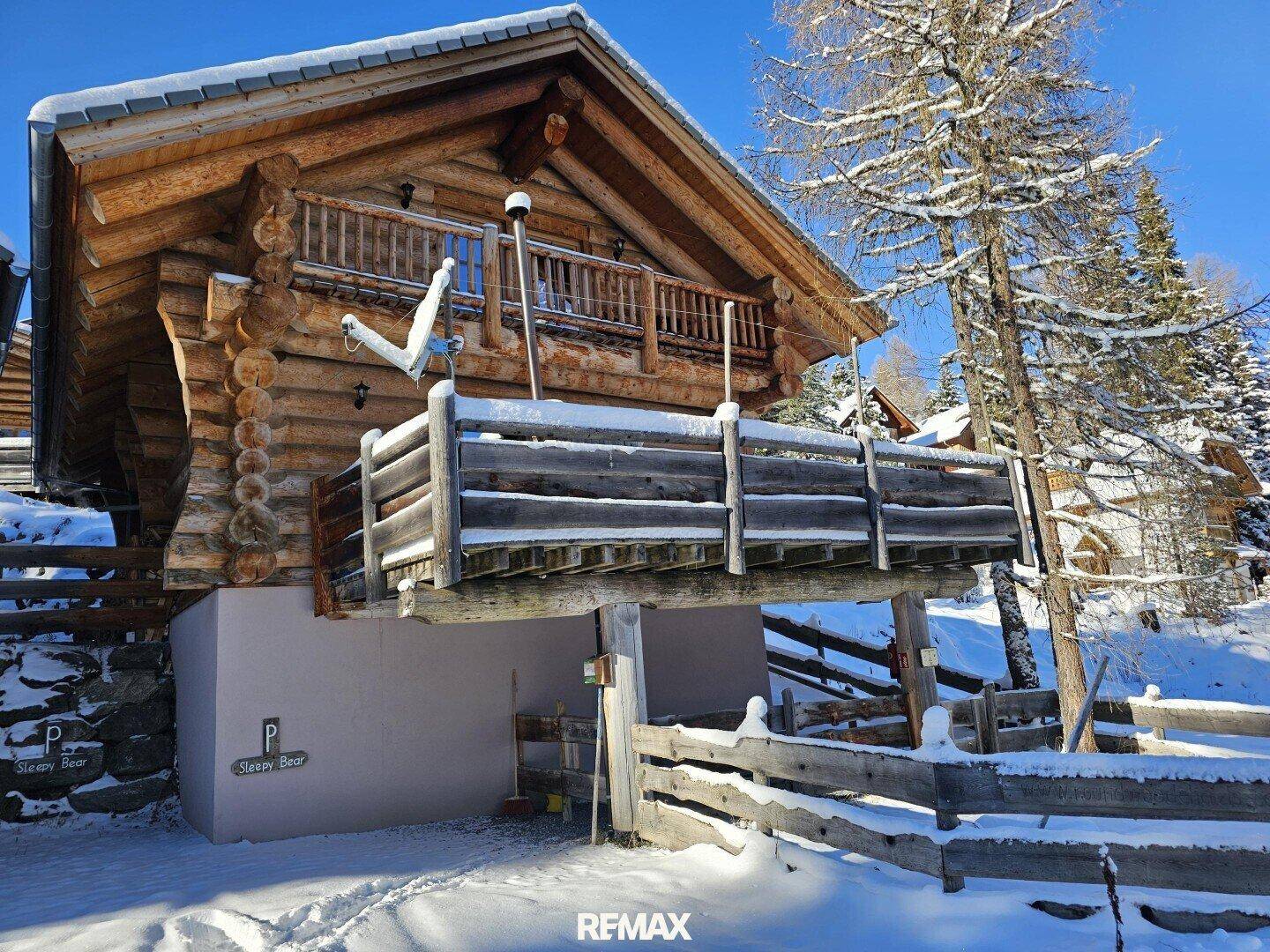 Holzchalet mit Balkon und einem überdachten Parkplatz darunter, alles schneebedeckt.