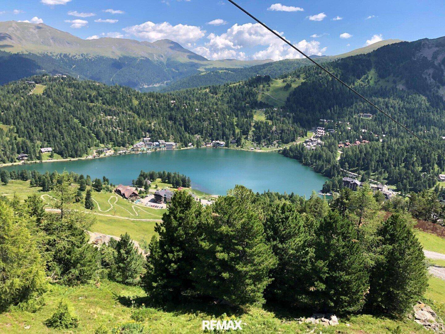 Malerischer Bergsee umgeben von bewaldeten Hängen und einem idyllischen Dorf unter blauem Himmel.