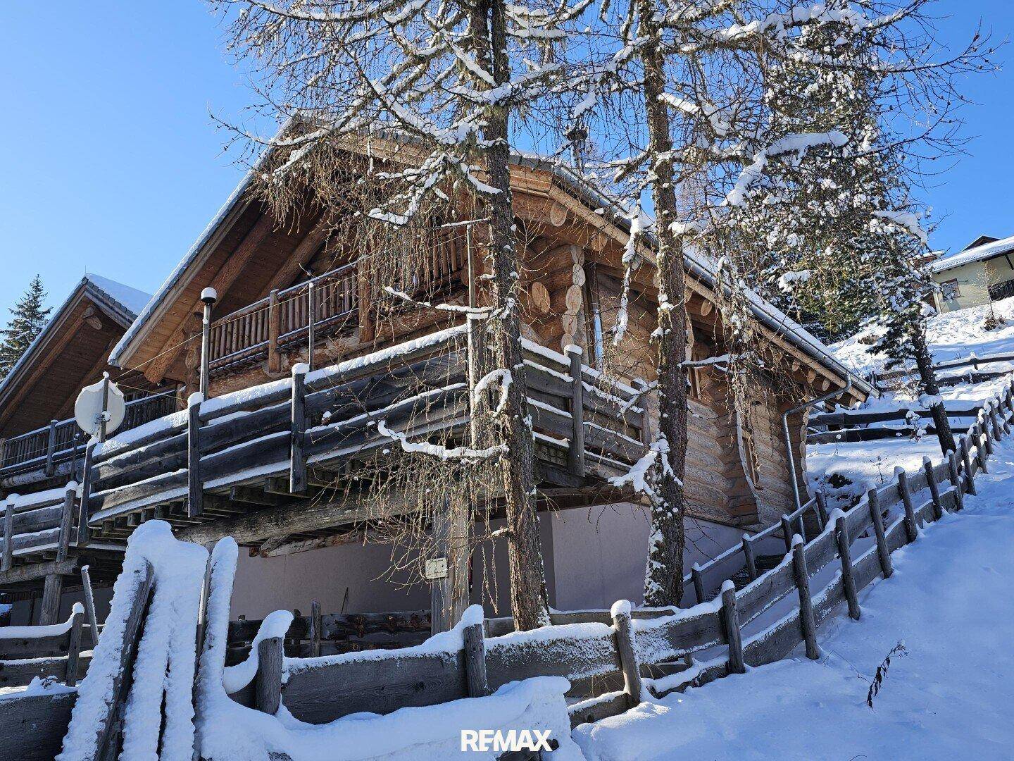 Blockhaus im Winter, mit Schnee bedeckt, umgeben von hohen Bäumen und einem rustikalen Holzzaun.
