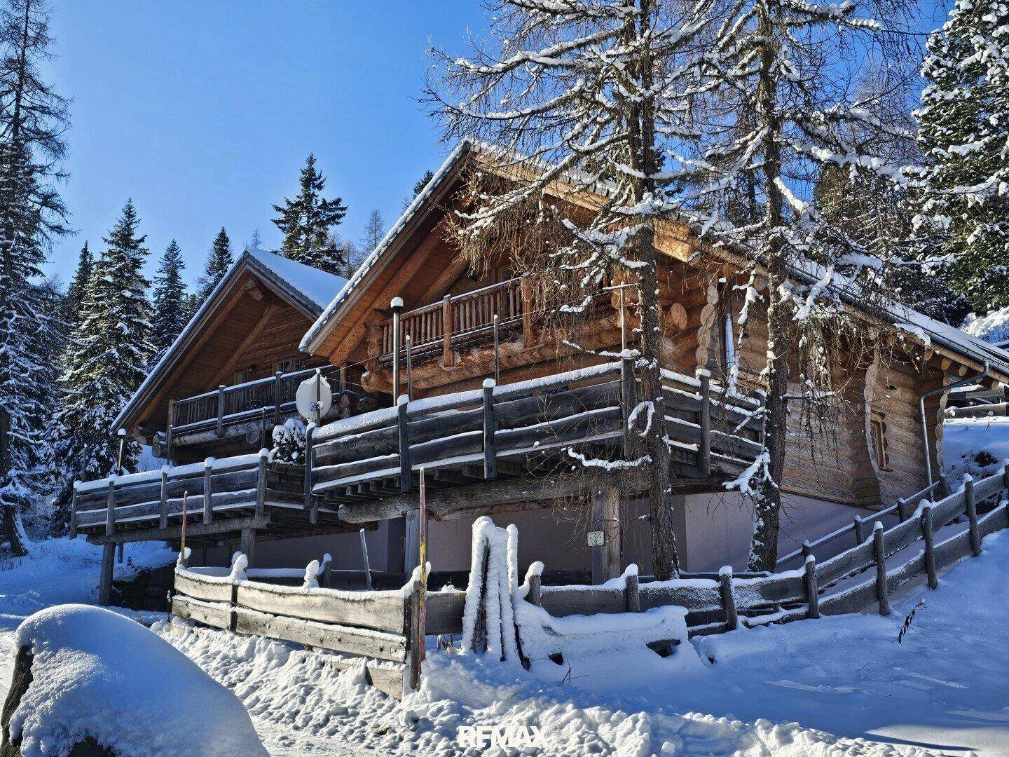 Verschneite Blockhäuser in winterlicher Landschaft, umgeben von Bäumen und einem Holzzaun.