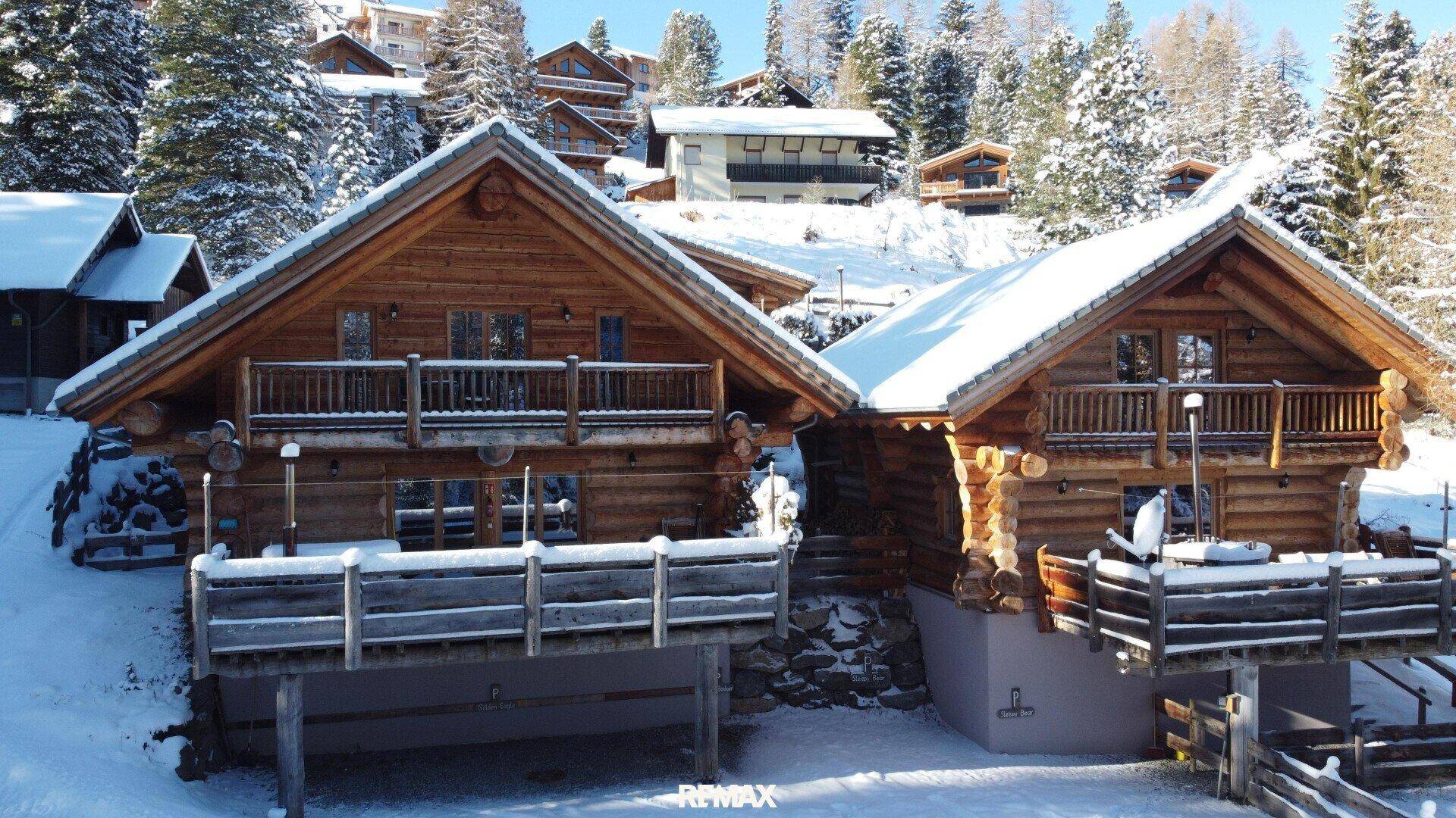 Zwei charmante Holzchalets in einer verschneiten Bergumgebung mit weiteren Gebäuden im Hintergrund.