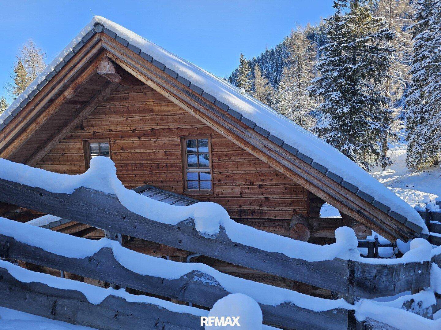 Seitliche Ansicht eines Holzchalets im Winter mit schneebedecktem Zaun und Bäumen.