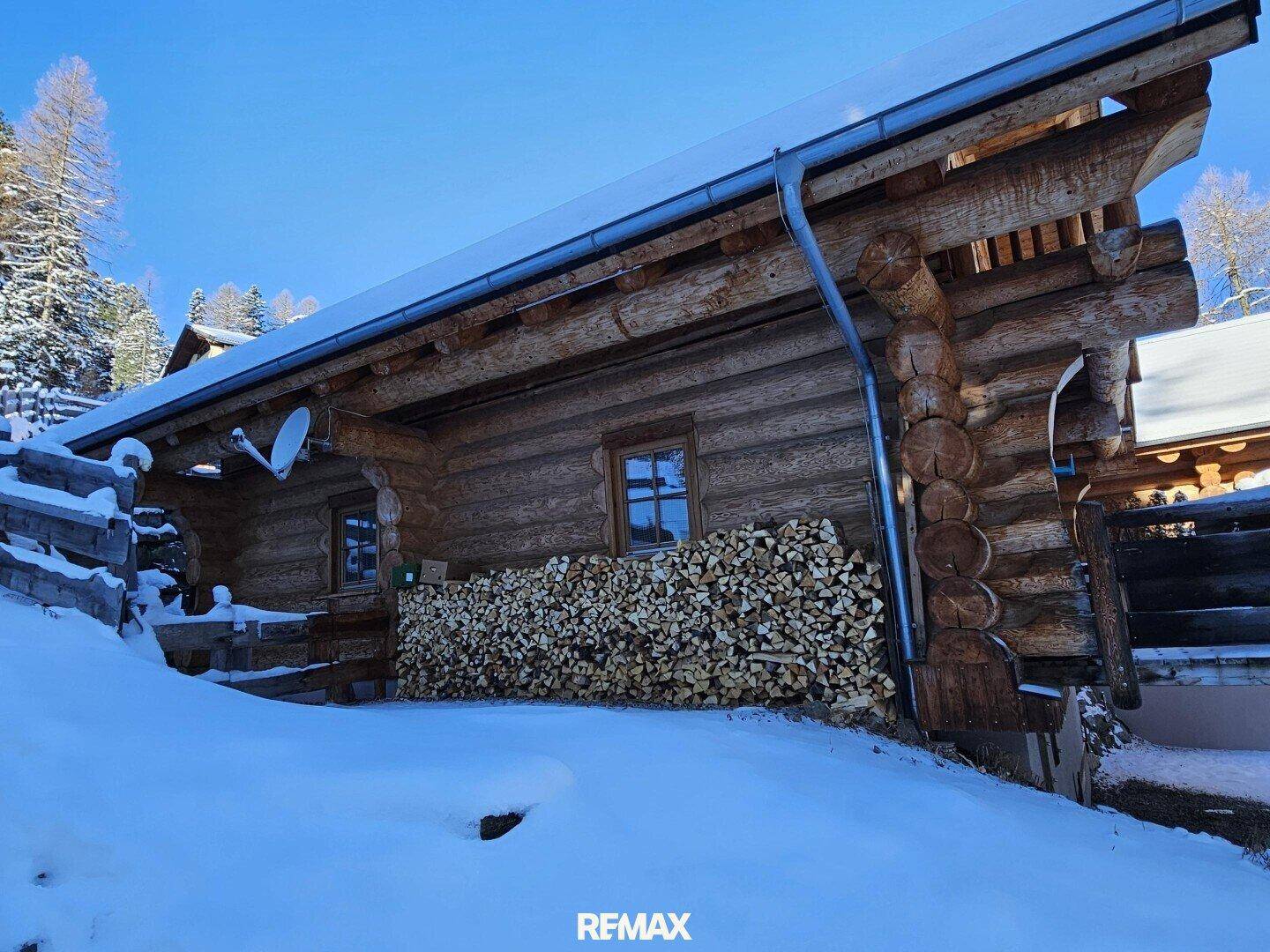Rustikales Holzhaus im Schnee mit gestapeltem Brennholz an der Fassade.