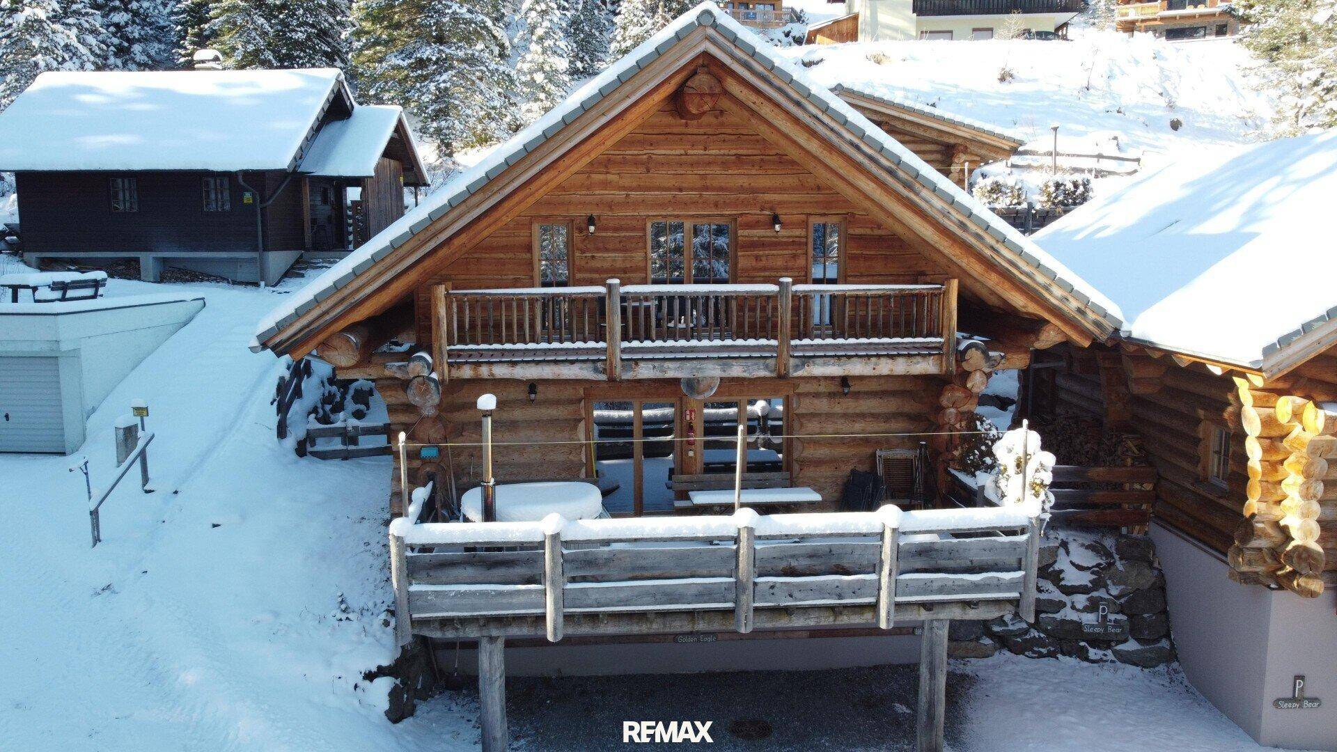 Holzchalet im Winter mit schneebedeckter Terrasse und Balkon, umgeben von verschneiten Bäumen.