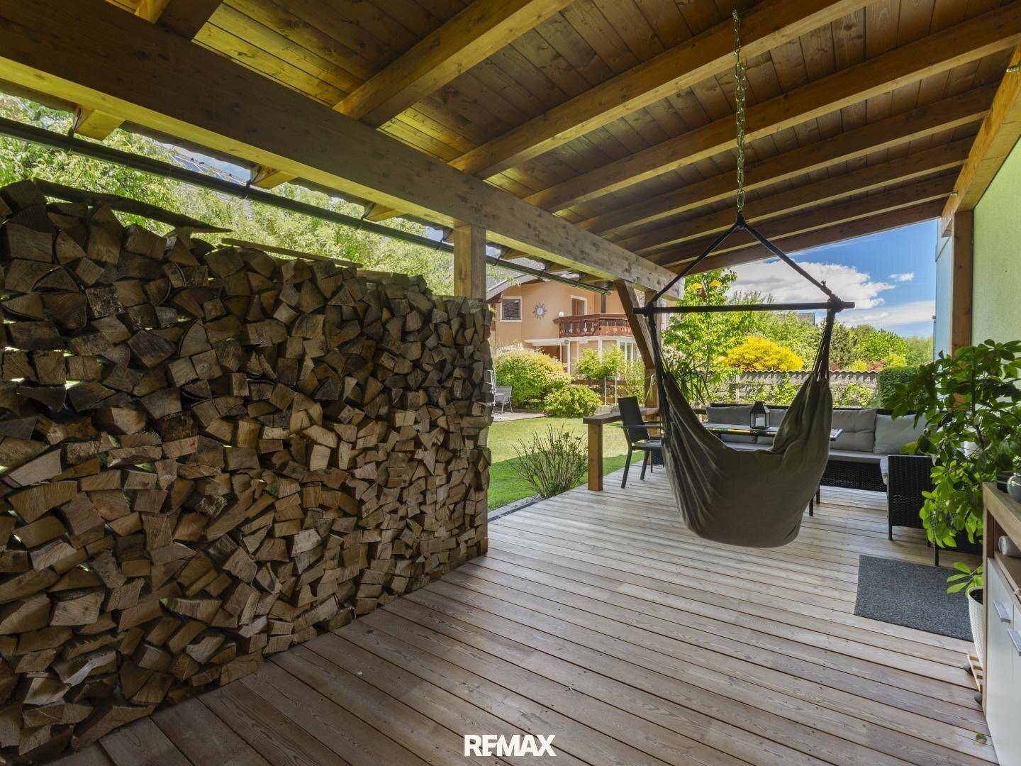 Überdachte Terrasse mit Holzdeck, Hängesessel und einem großen Stapel Brennholz als Sichtschutz.