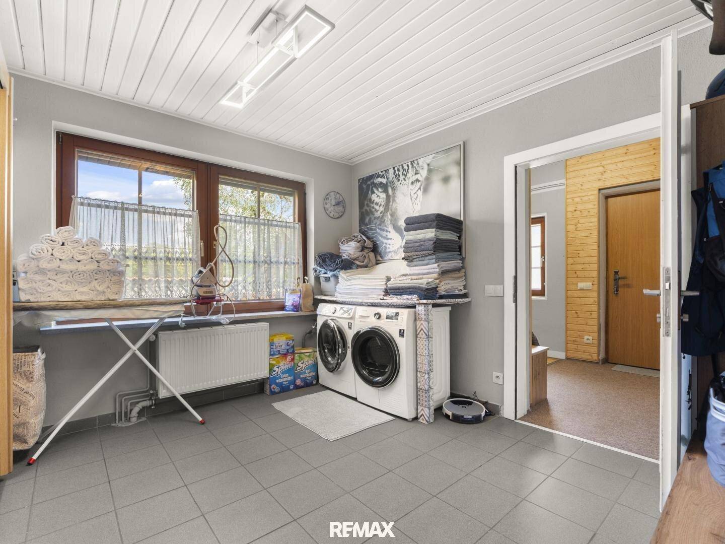 Praktischer Waschraum mit Waschmaschine, Trockner und Bügelbrett, ausgestattet mit Fliesenboden und Fenster.