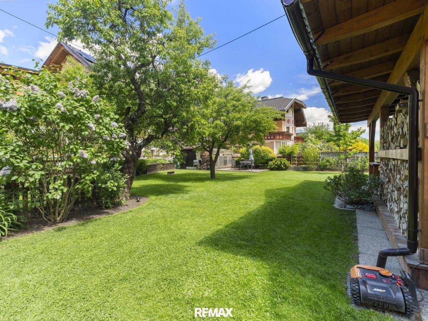 Gepflegter Rasenbereich im Garten mit Bäumen, Sträuchern und einem Mähroboter unter blauem Himmel.