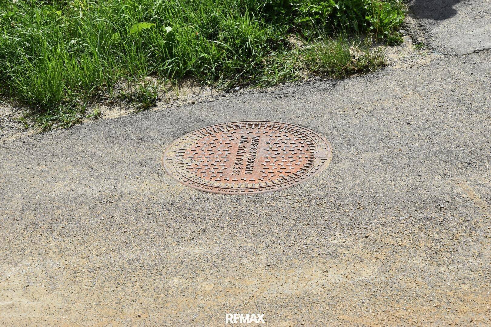 Runder Kanaldeckel aus Metall auf einer asphaltierten Straße, umgeben von dunklem Asphalt.
