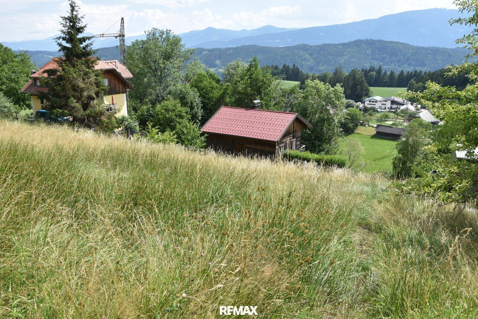 Weitläufige Wiese mit Blick auf traditionelle Häuser und eine malerische Berglandschaft in der Ferne.