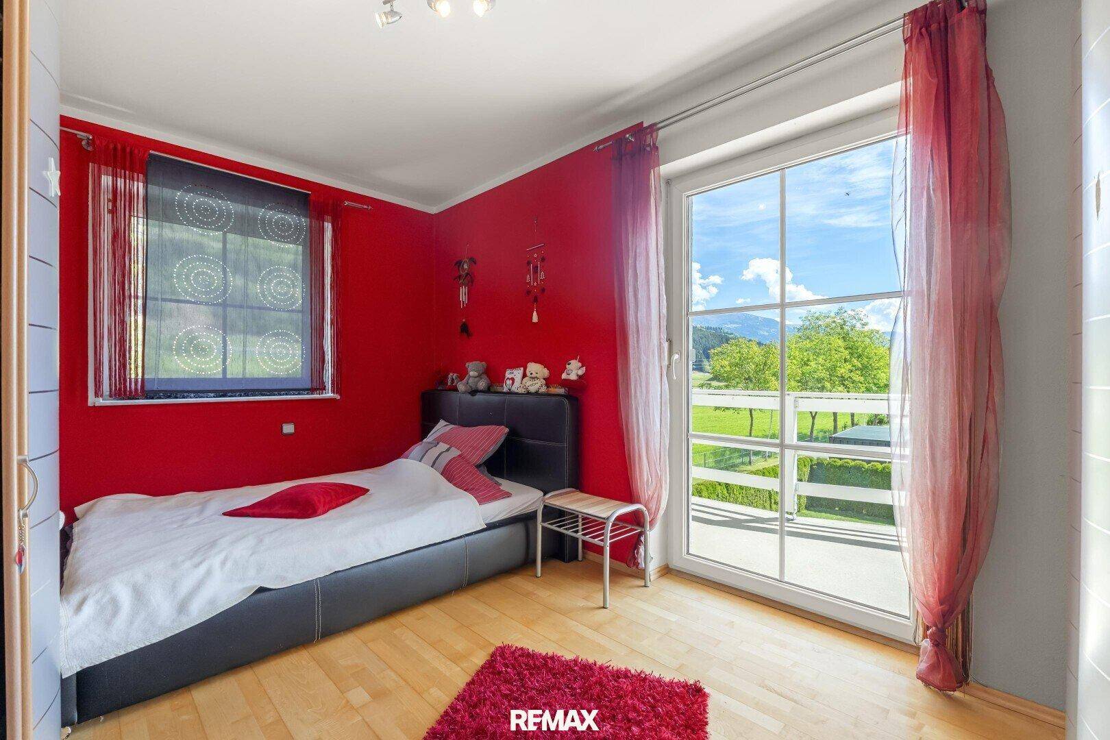 Farbiges Schlafzimmer mit roter Wand, Holzboden und großem Fenster mit Blick ins Grüne.