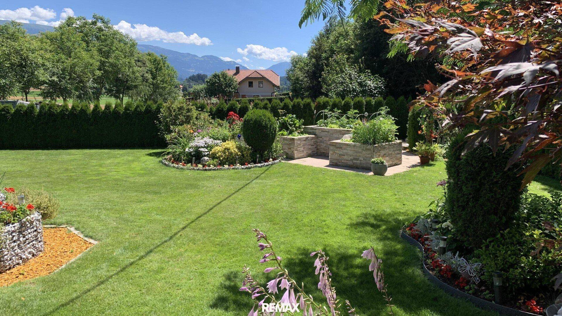 Gepflegter Garten mit Rasenflächen, Blumenbeeten und Hochbeeten vor Bergkulisse.