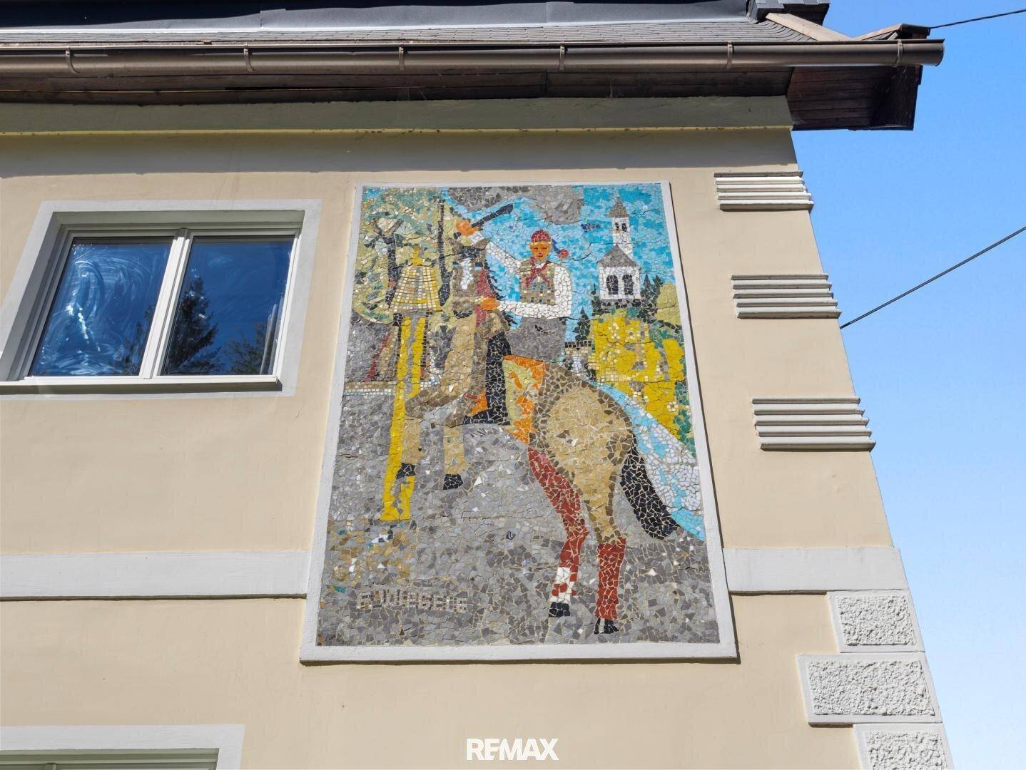 Nahaufnahme eines farbenfrohen Mosaiks an der hellen Fassade des Hauses unter blauem Himmel.