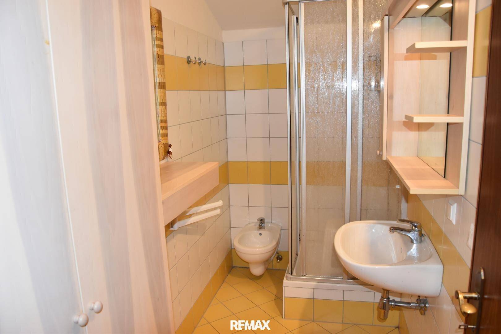 Funktionales Badezimmer mit gelb-weißen Fliesen, Dusche, Bidet und Waschbecken.