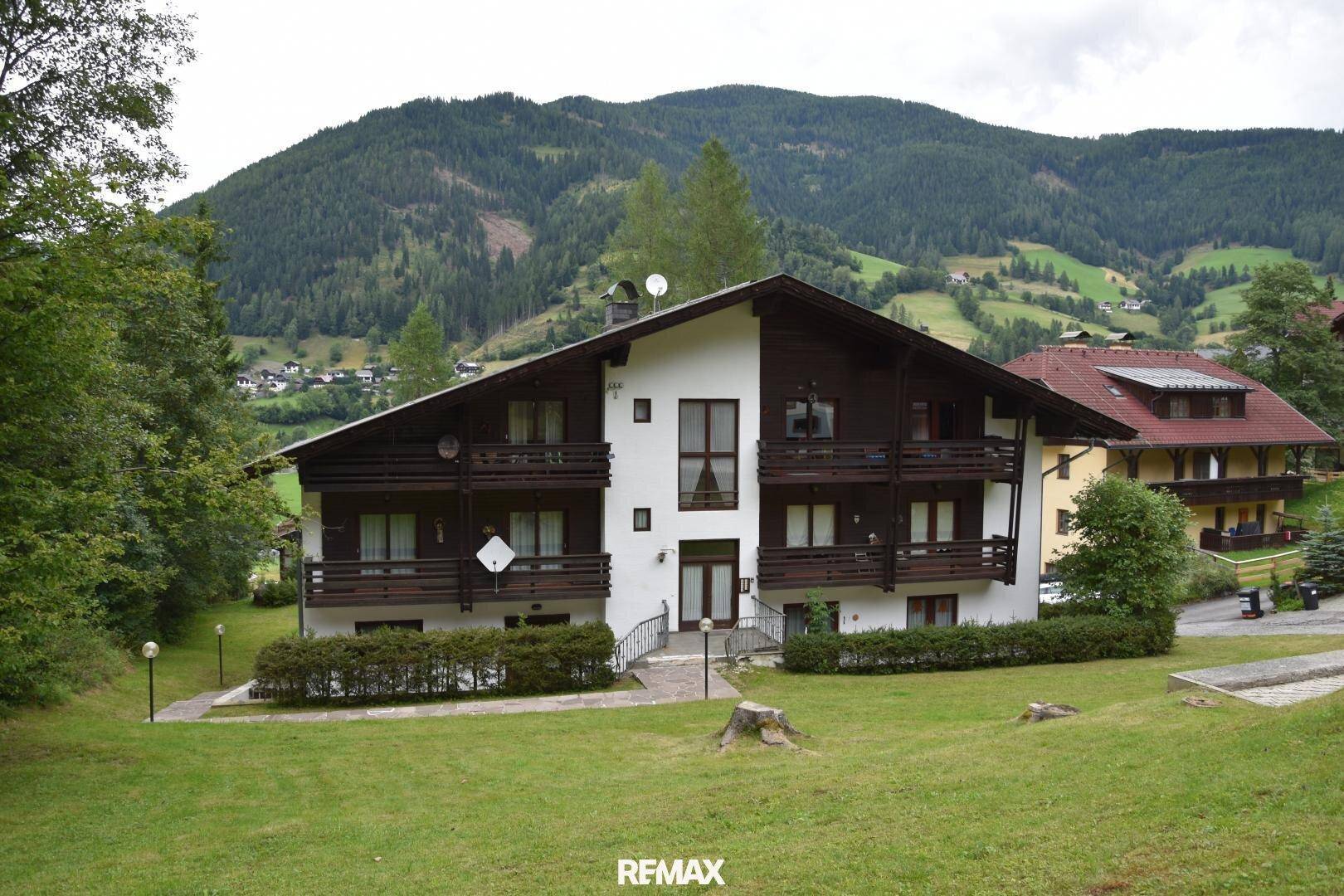 Alpines Mehrfamilienhaus mit dunklen Holzdetails und Balkonen vor bergiger Kulisse.