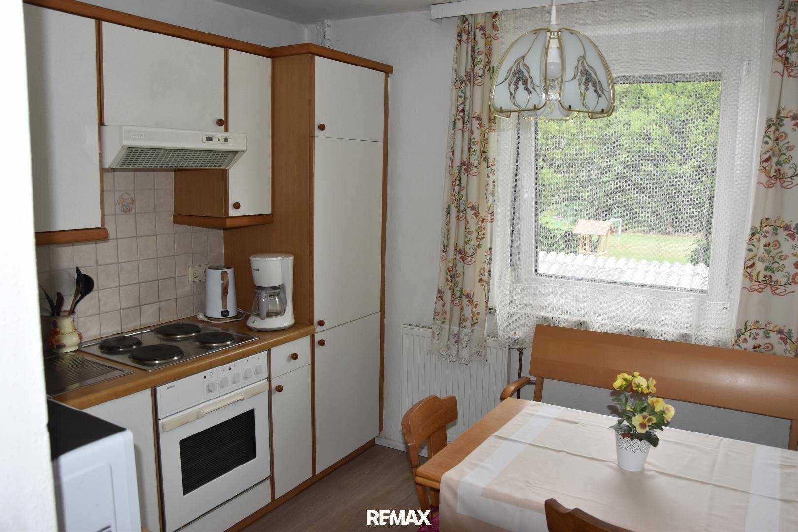 Küche mit weißen und holzfarbenen Schränken, Elektroherd und Essbereich am Fenster mit Grünblick.