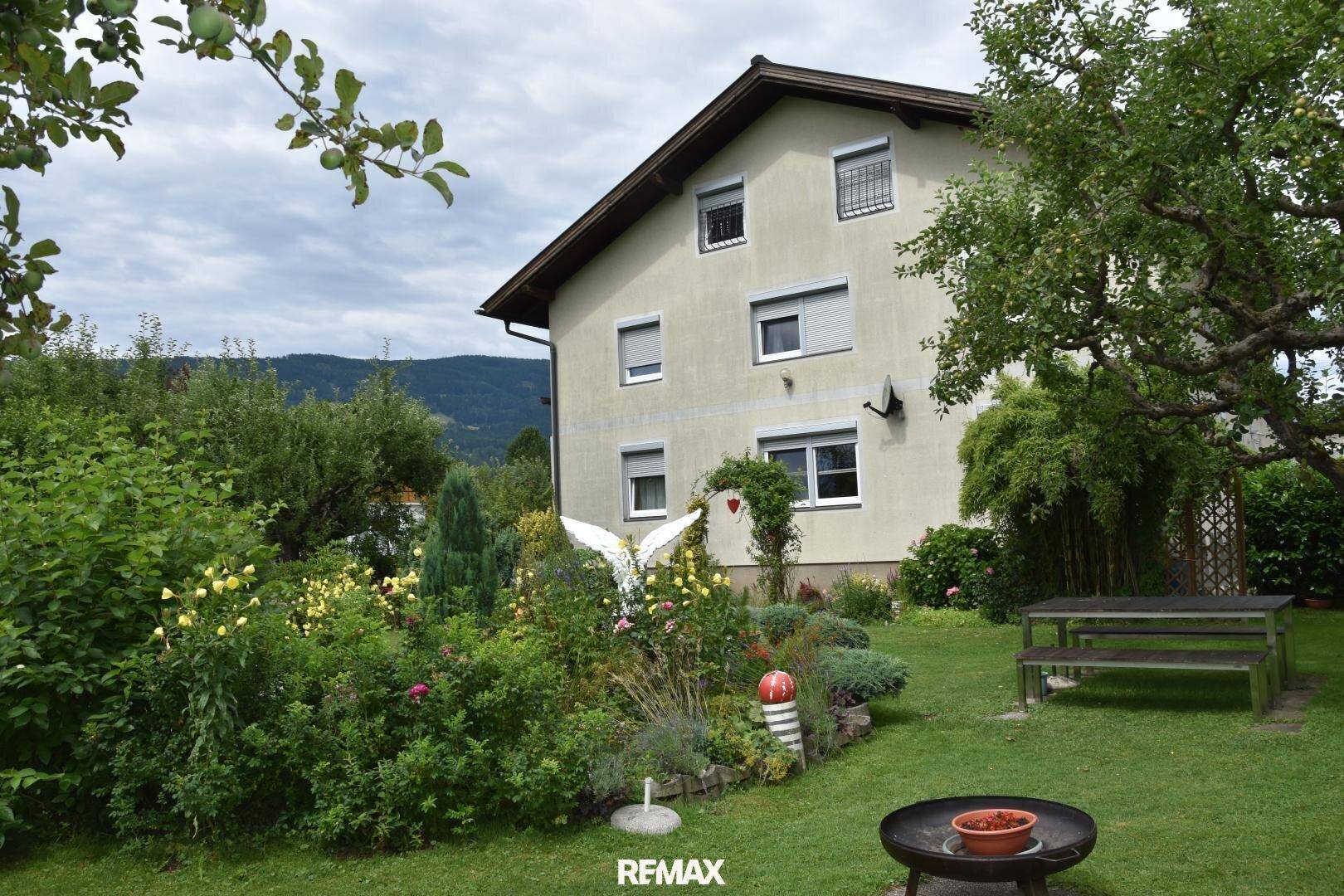 Rückansicht des Hauses mit Garten, Blumenbeeten, einer Holzbank und einer Feuerschale im Vordergrund.