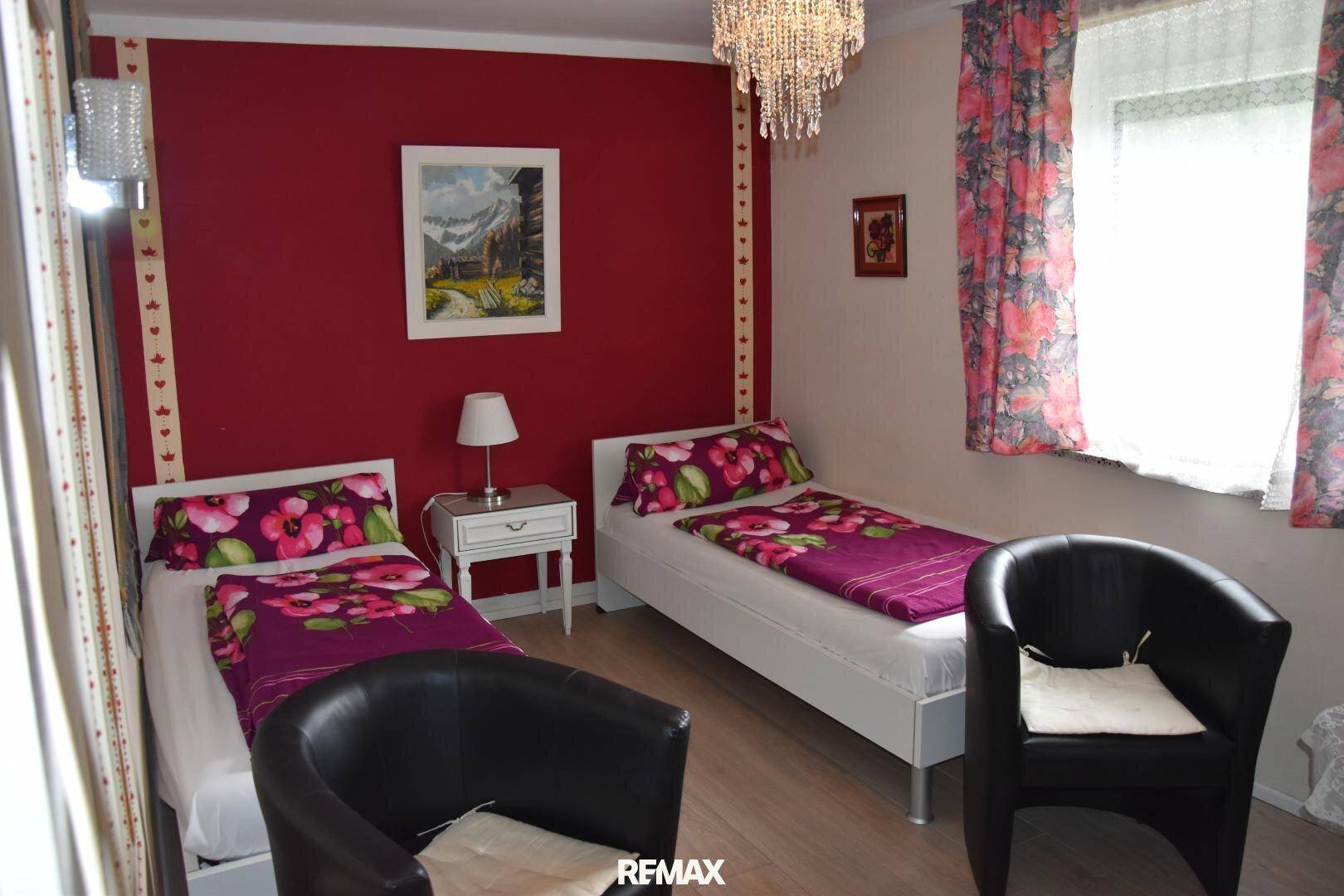 Schlafzimmer mit roten Wänden, zwei Einzelbetten und einem Kronleuchter, mit Blick nach draußen.