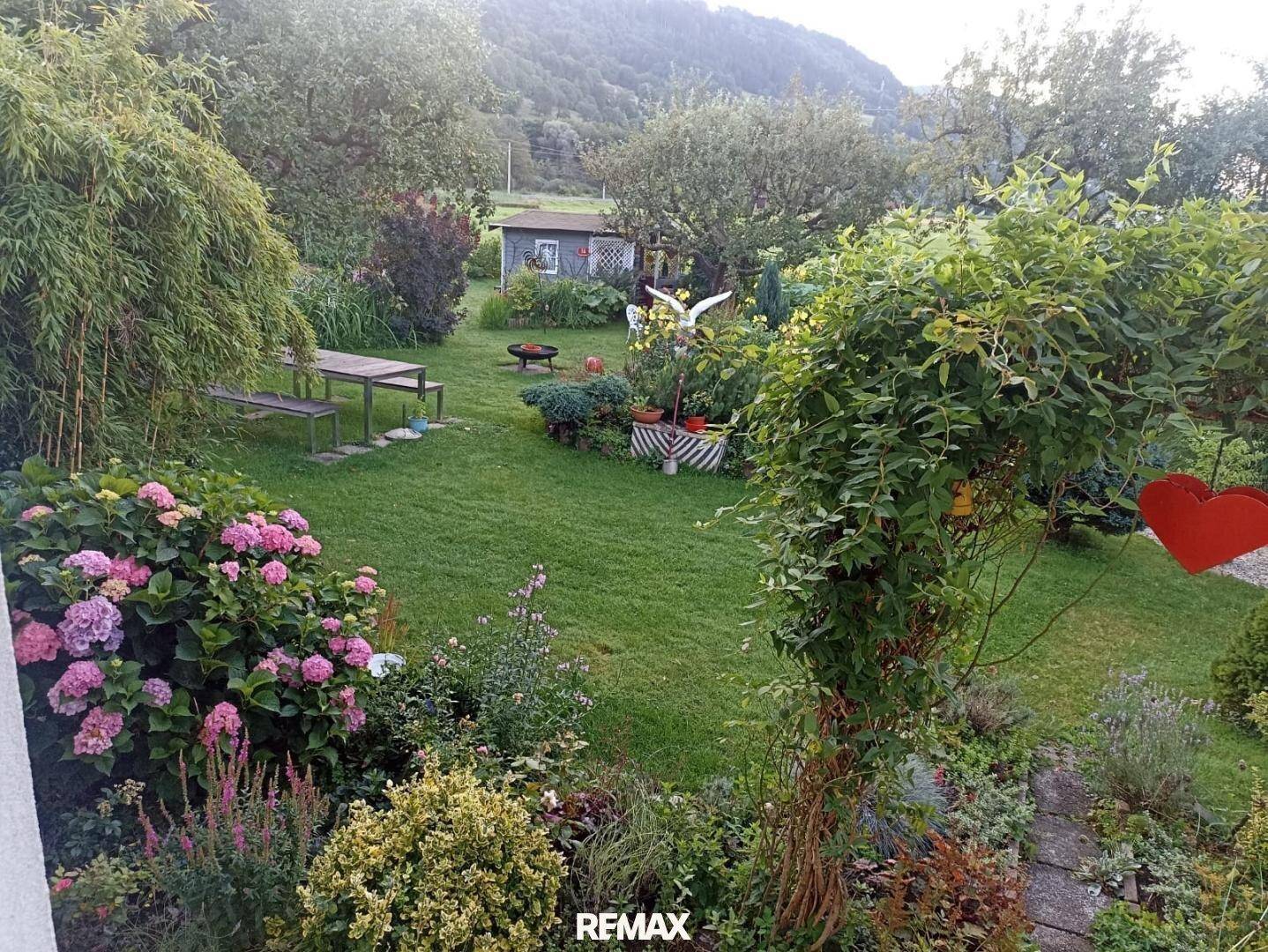 Großer, blühender Garten mit verschiedenen Pflanzen, Rasen und einem Esstisch im Freien.